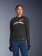 Mujer con chaqueta Alpinestars Stella SMX Air, negro, blanco y rojo flúor, chaqueta de moto textil con malla ventilada y logotipo blanco en el pecho, combinada con vaqueros azules y guantes de moto negros, ajuste femenino para conducción deportiva