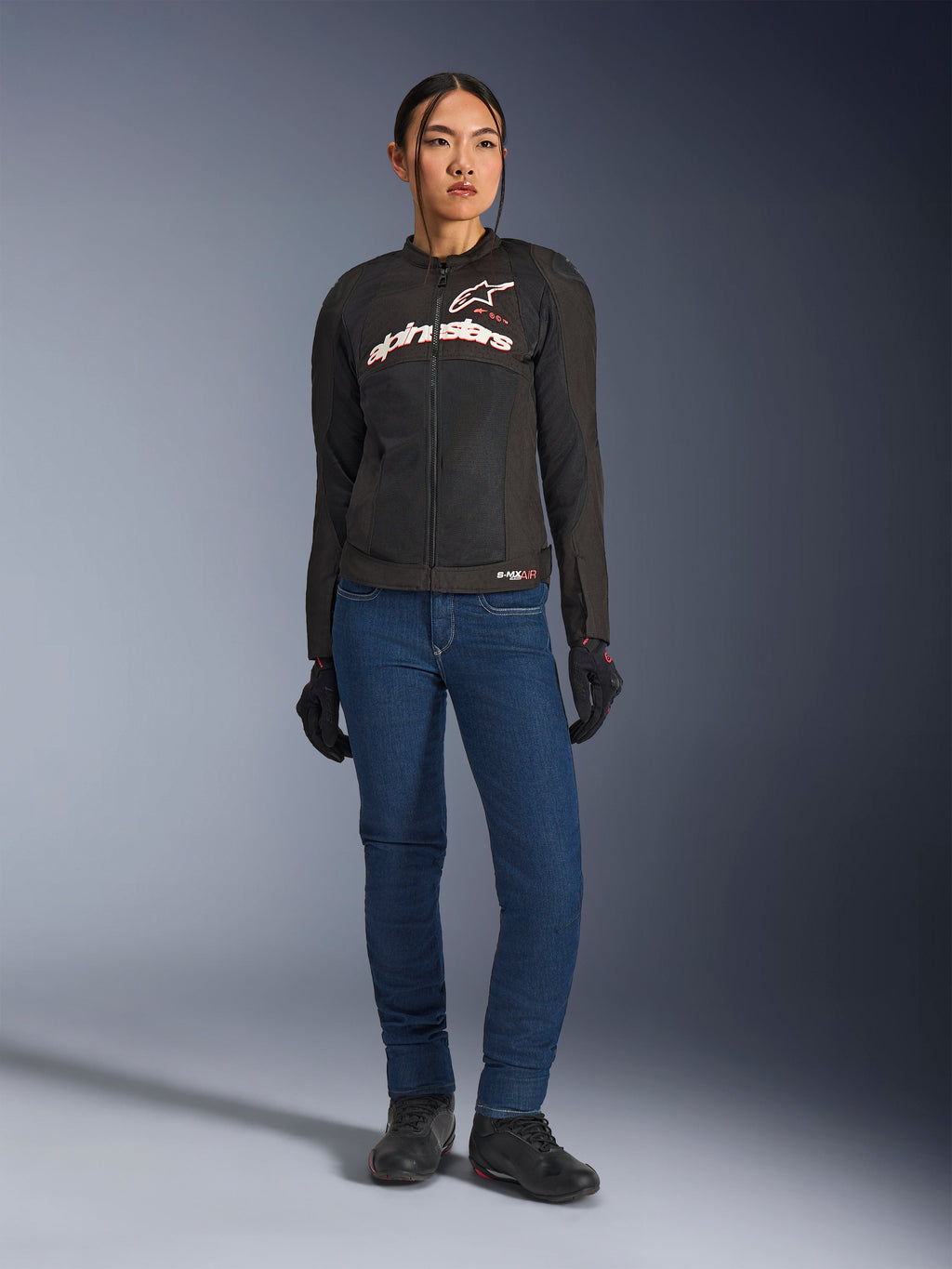 Piloto femenina con la chaqueta Alpinestars Stella SMX Air, negra con logotipos en blanco y rojo flúor, combinada con pantalones vaqueros azules y guantes negros, chaqueta textil deportiva ventilada para conducción en carretera