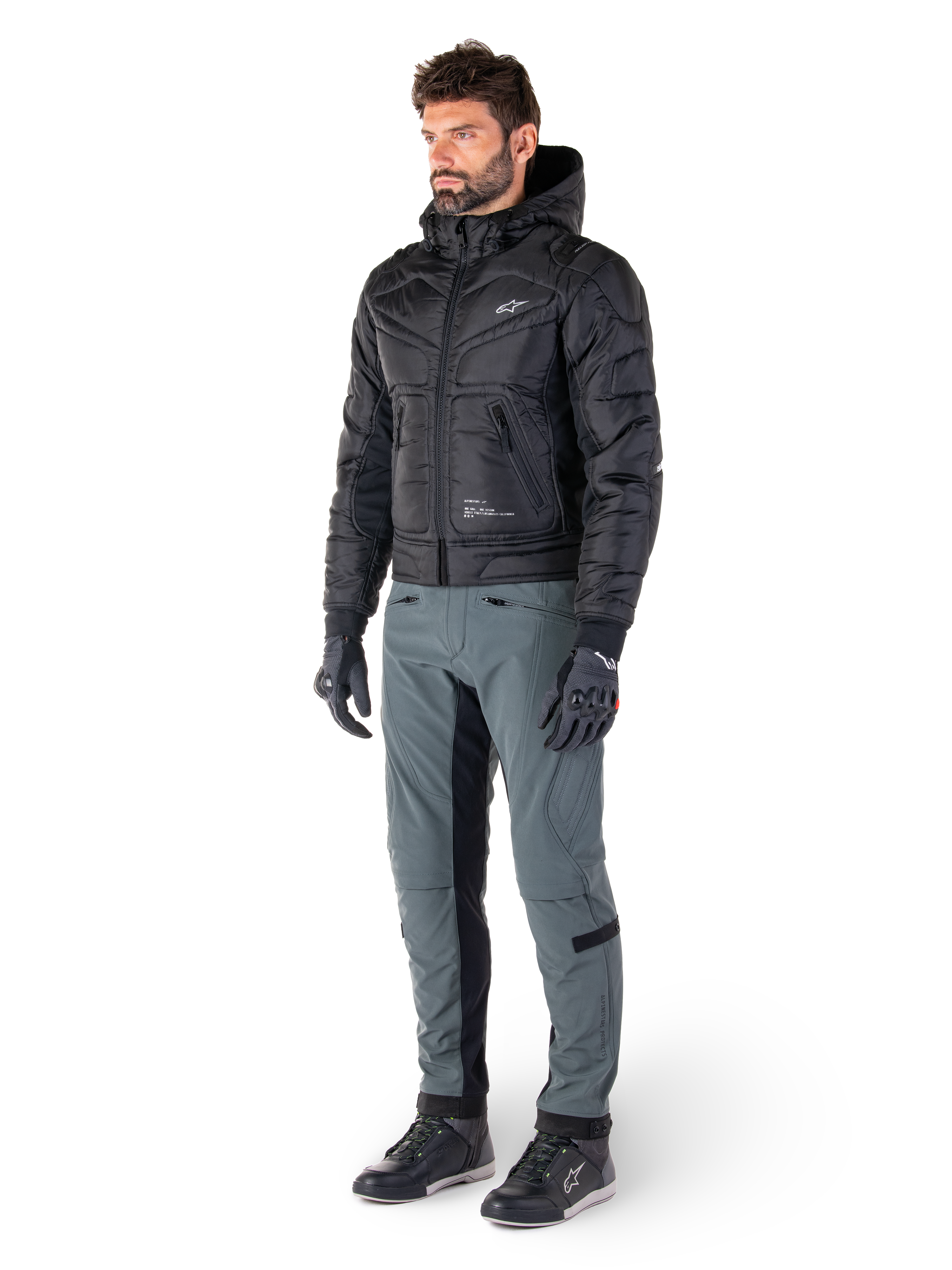 Pantalones Alpinestars Alden en Urban Gray, llevados por un modelo con una chaqueta negra acolchada con capucha, guantes y calzado de conducción, con detalles acolchados en los laterales y paneles interiores negros en las piernas, equipamiento urbano para moto inspirado en el estilo streetwear.