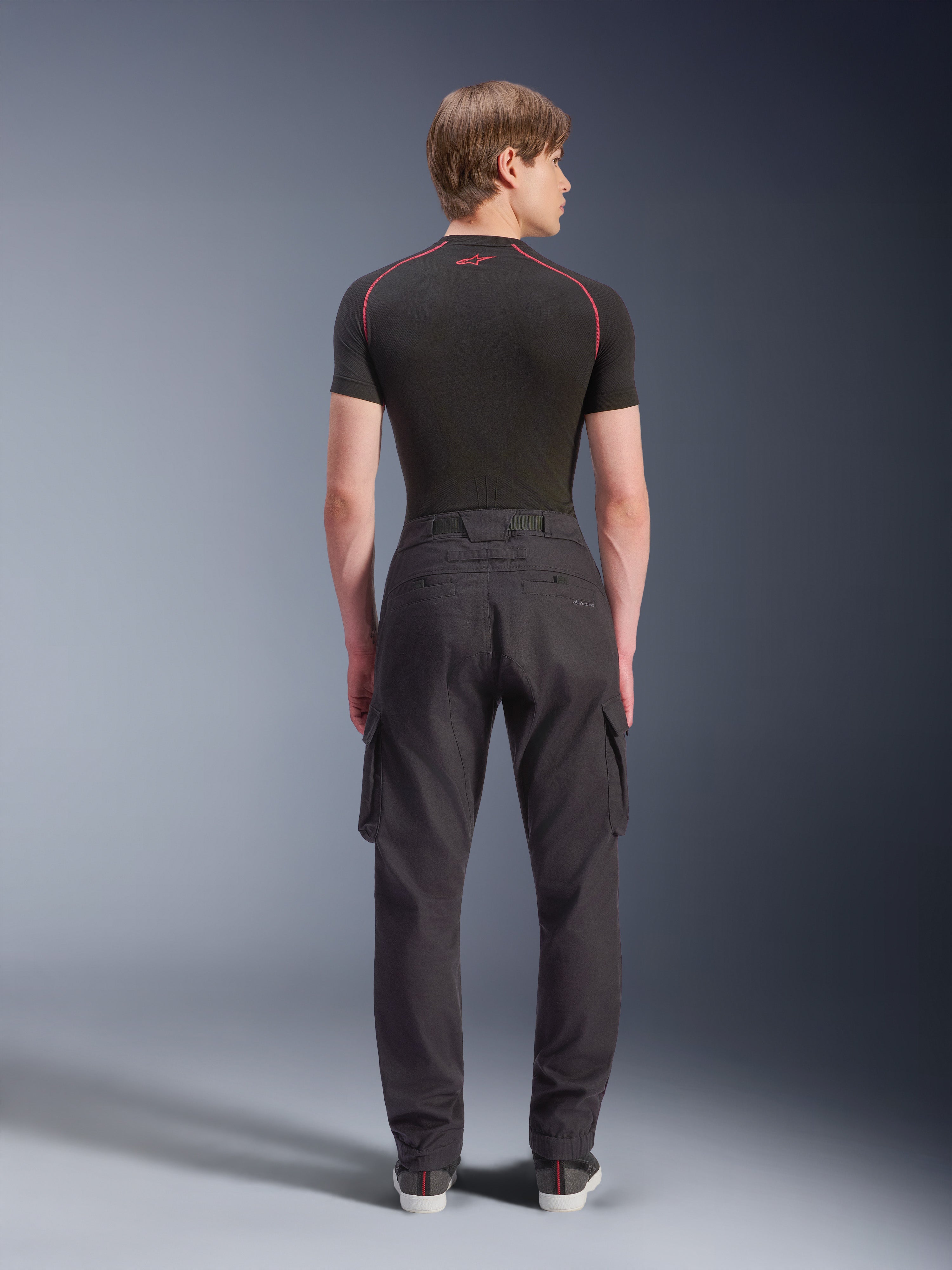 Pantalones Cargo Alpinestars Flex-AST, pantalones cargo para moto, negros, vista trasera en modelo, con un corte regular ahusado con dobladillos elásticos y bolsillos cargo laterales, combinados con una capa base negra y zapatillas casuales