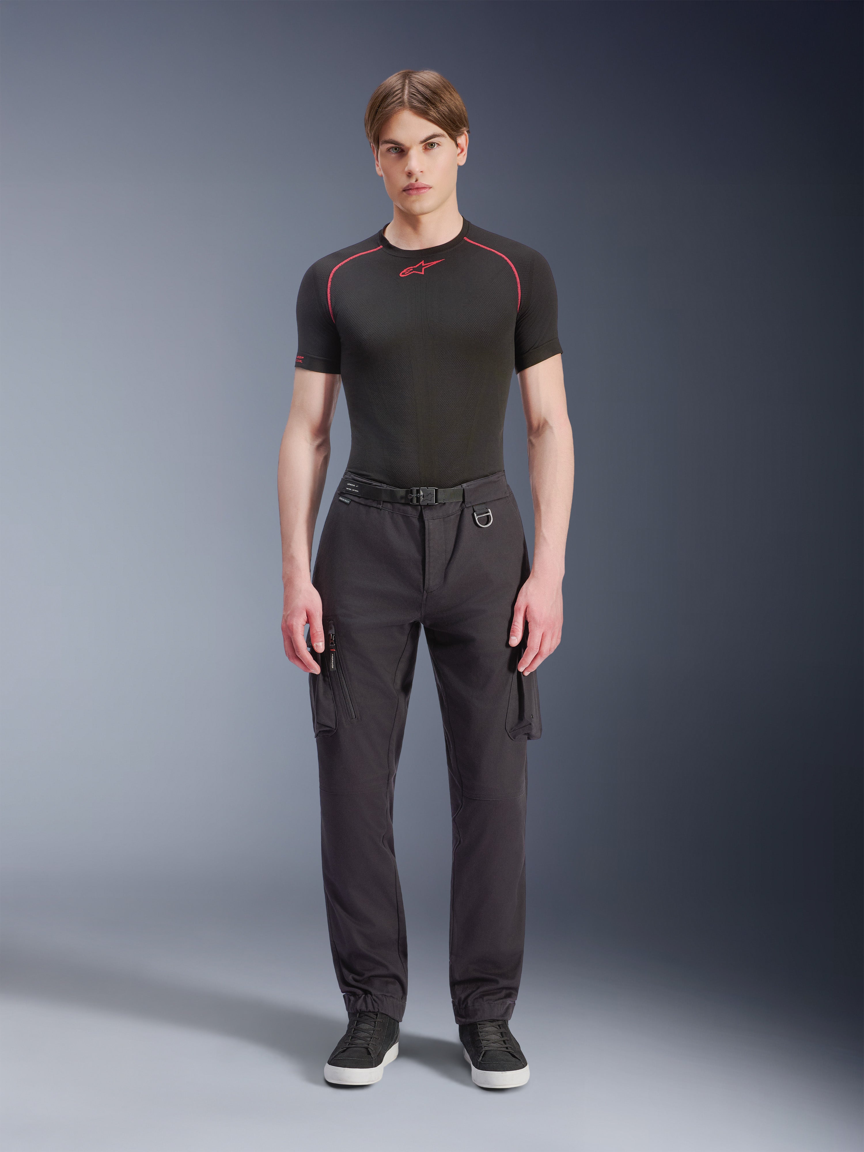 Pantalones Cargo Alpinestars Flex-AST, negros, pantalones de moto urbanos llevados por un modelo masculino con una parte superior negra de manga corta y zapatillas, con un ajuste regular ahusado, bolsillos cargo y detalle de hebilla de cinturón integrada