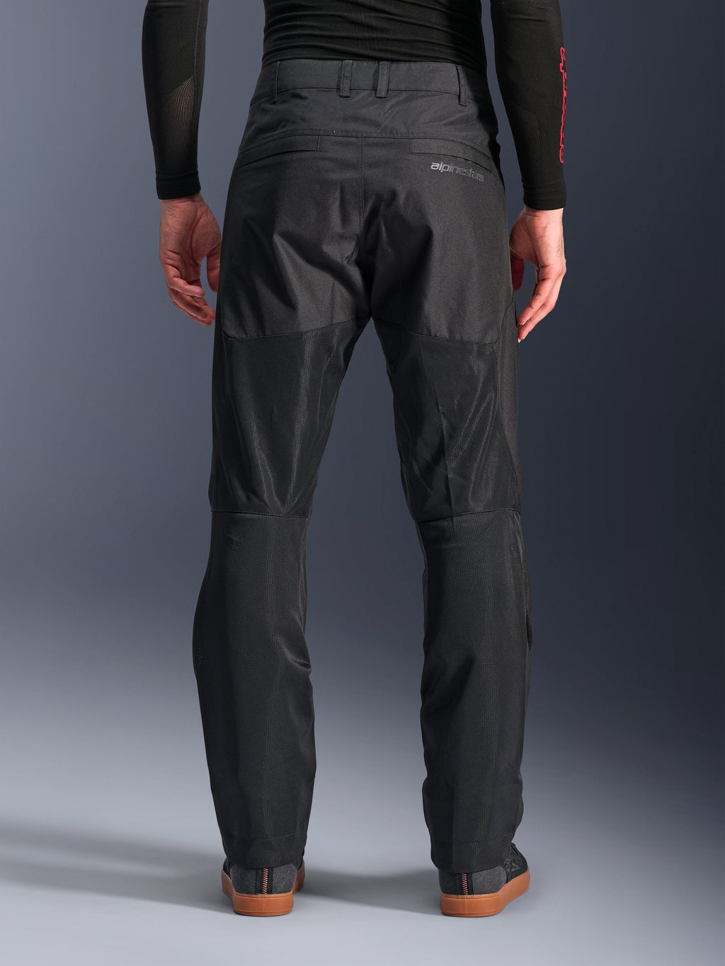 Pantalones Alpinestars C-1 Air, vista trasera, negros, pantalones urbanos de moto fabricados en tejido textil con amplios paneles de malla técnica en la parte posterior de las piernas para mayor ventilación, diseño limpio con marca sutil, combinados con una capa base negra y calzado casual para conducir.