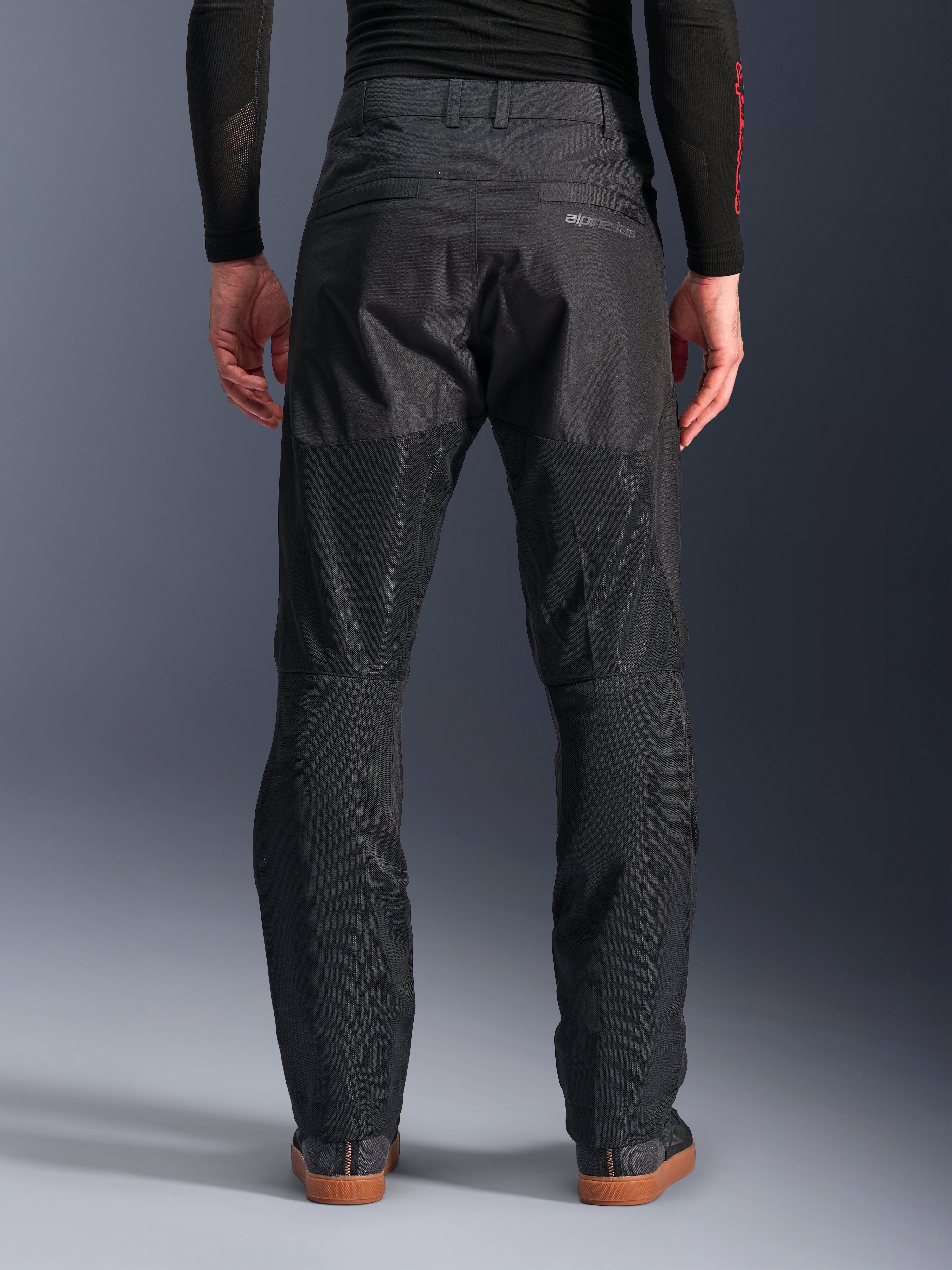 Pantalones Alpinestars C-1 Air, vista trasera, negros, pantalones urbanos de moto fabricados en tejido textil con amplios paneles de malla técnica en la parte posterior de las piernas para mayor ventilación, diseño limpio con marca sutil, combinados con una capa base negra y calzado casual para conducir.