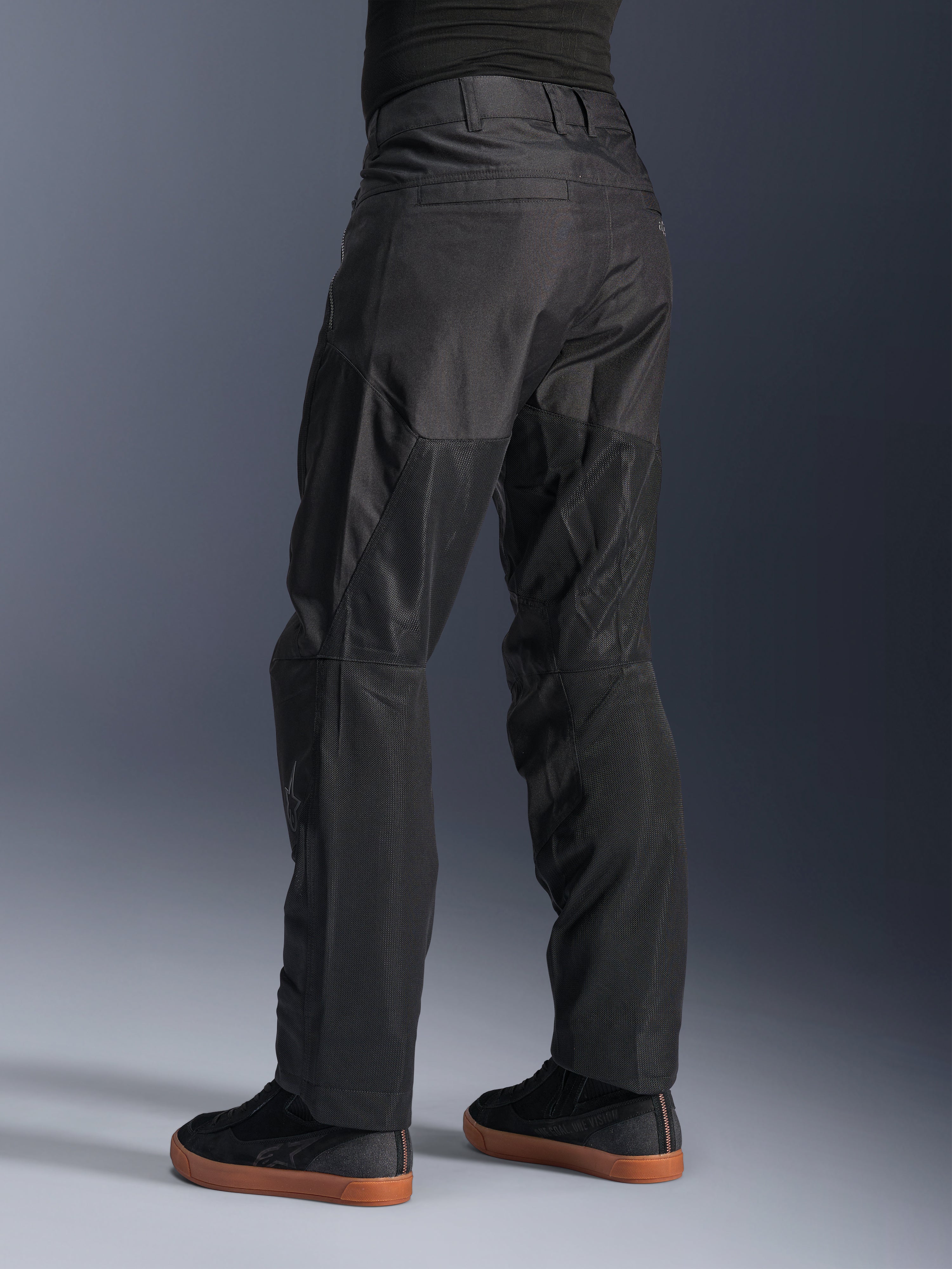 Alpinestars C-1 Air Pants, pantalones urbanos de moto, color negro, vista trasera que muestra grandes paneles de malla técnica en la parte posterior de las piernas para ventilación, estilo limpio para conducción urbana, combinados con zapatillas casuales negras.