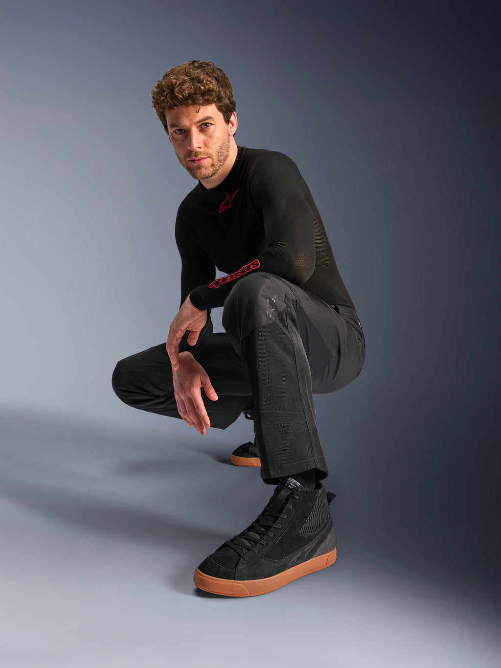 Pantalones Alpinestars C-1 Air, pantalones de moto urbanos, negros, modelo agachado en un estudio, combinados con una camiseta negra de manga larga y zapatillas negras con suela color canela, con paneles de malla transpirable y un estilo urbano contemporáneo