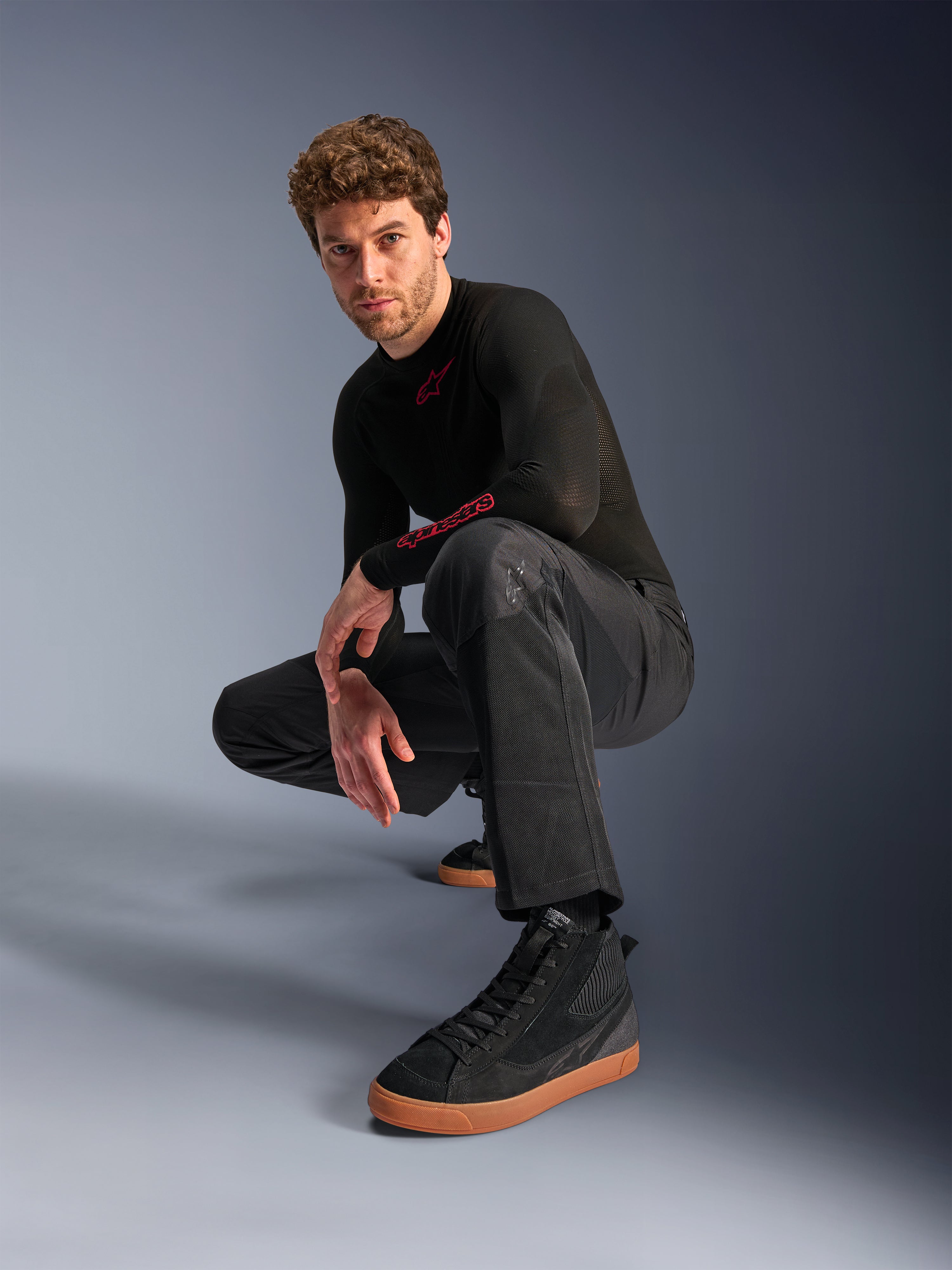Pantalones Alpinestars C-1 Air, pantalones de moto urbanos, negros, modelo agachado en un estudio, combinados con una camiseta negra de manga larga y zapatillas negras con suela color canela, con paneles de malla transpirable y un estilo urbano contemporáneo