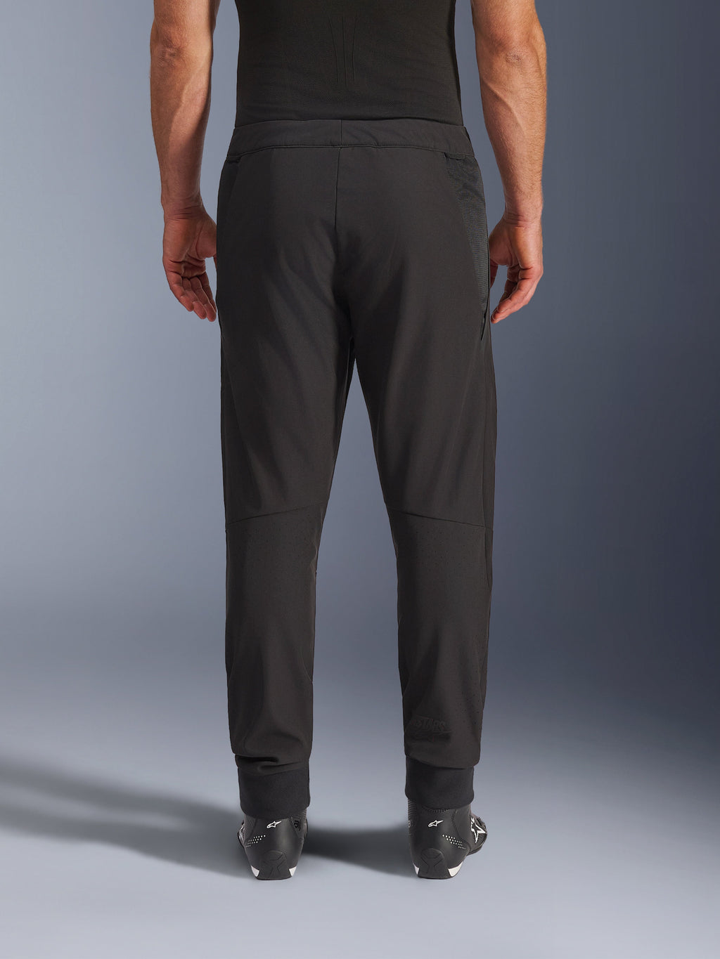 Pantalones Alpinestars Aeroshell Airflow, negros, vista trasera, con zonas perforadas por láser en las pantorrillas y paneles de malla, usados con botas Alpinestars negras, diseño de softshell ligero para deporte y turismo