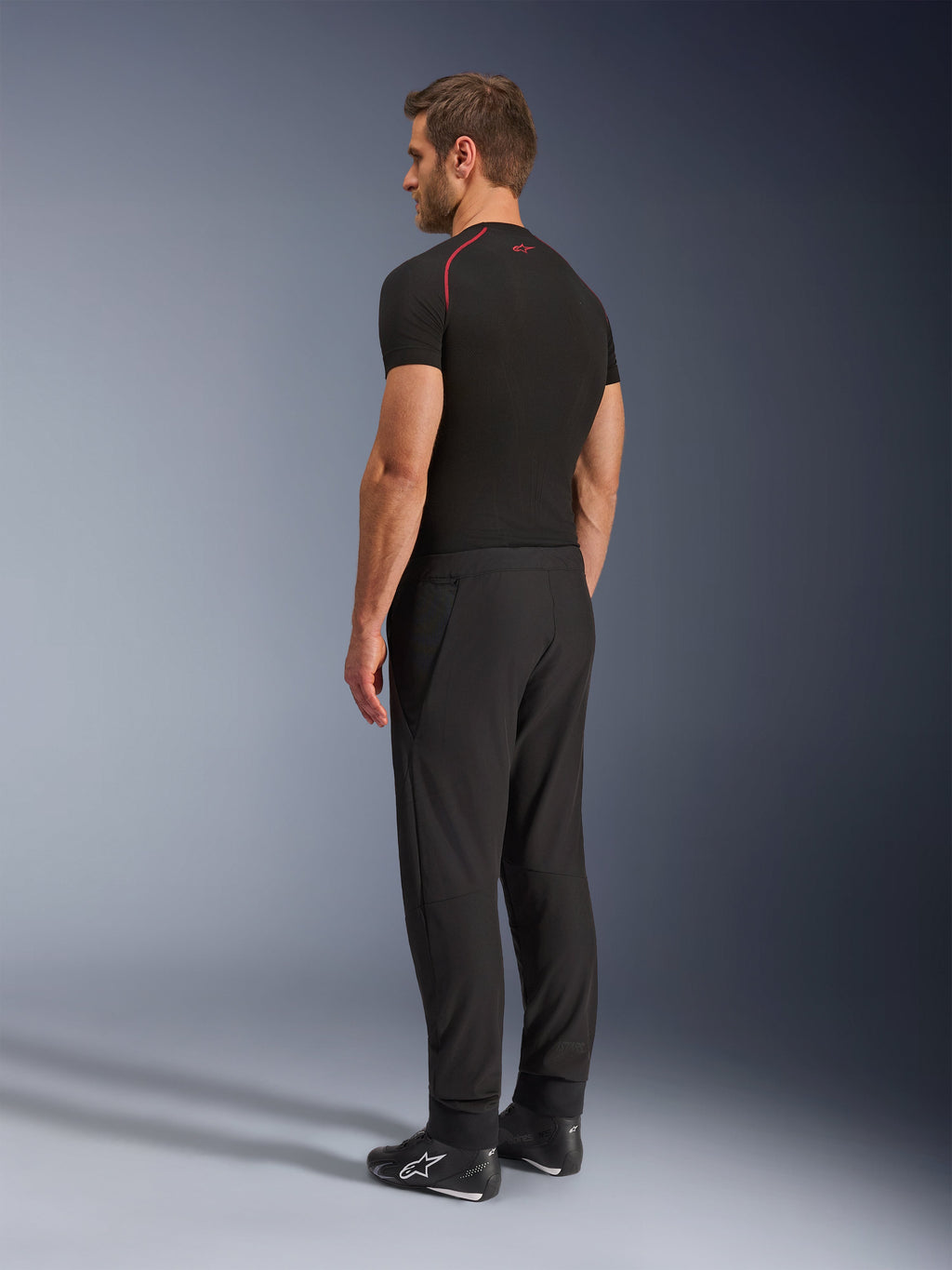 Pantalones Alpinestars Aeroshell Airflow, pantalones de moto negros, tejido softshell ligero, usados con una camiseta técnica negra de manga corta y botas de conducción negras, vista de perfil lateral trasera que muestra el corte ajustado y la construcción ergonómica.