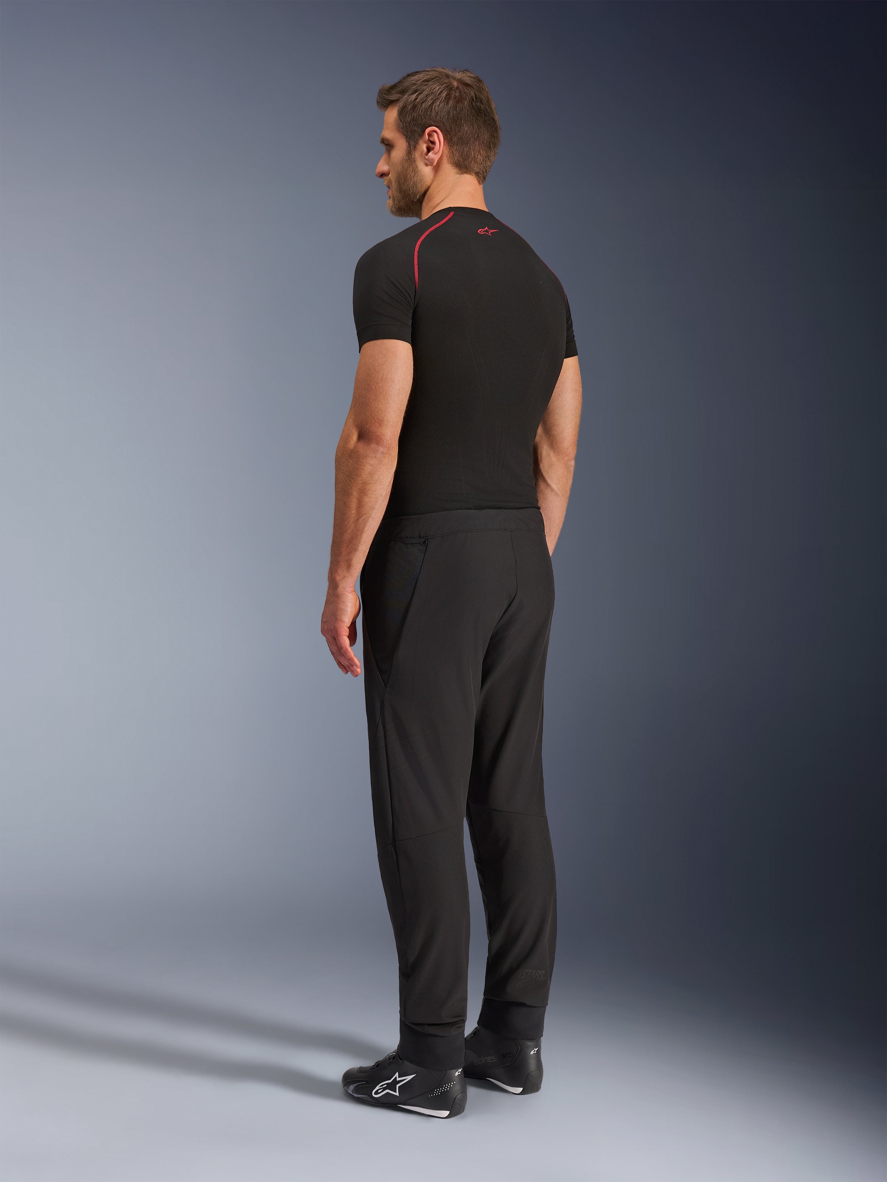 Pantalones Alpinestars Aeroshell Airflow, pantalones de moto negros, tejido softshell ligero, usados con una camiseta técnica negra de manga corta y botas de conducción negras, vista de perfil lateral trasera que muestra el corte ajustado y la construcción ergonómica.