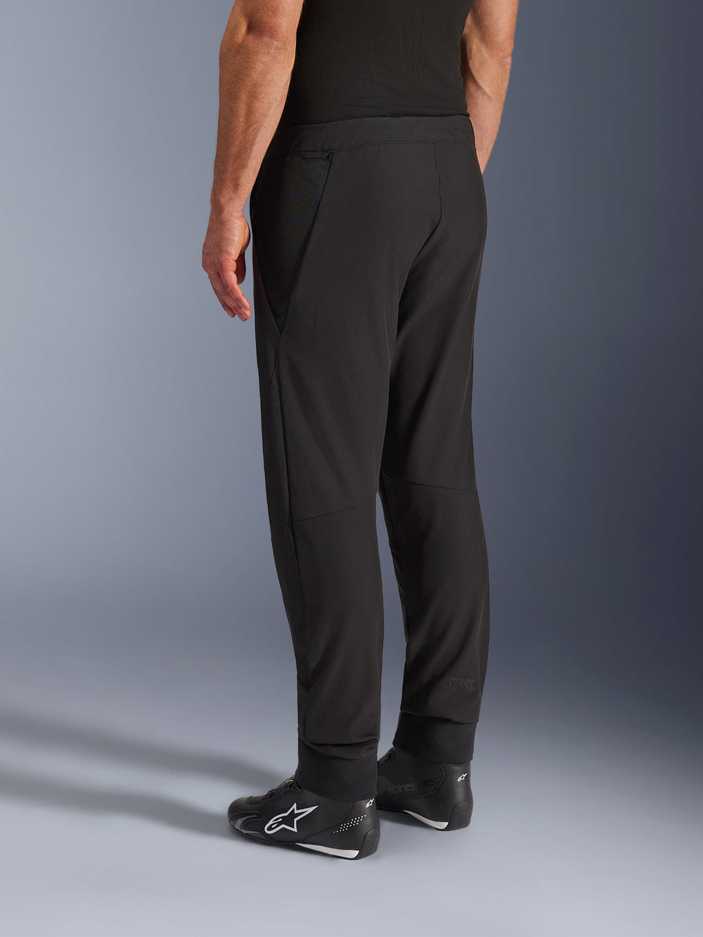 Alpinestars Aeroshell Airflow Pants, pantalones de moto negros, vista trasera en tres cuartos, construcción de softshell elástico ligero con paneles de malla integrados, puños de tobillo ajustables, usados con calzado de conducción Alpinestars negro