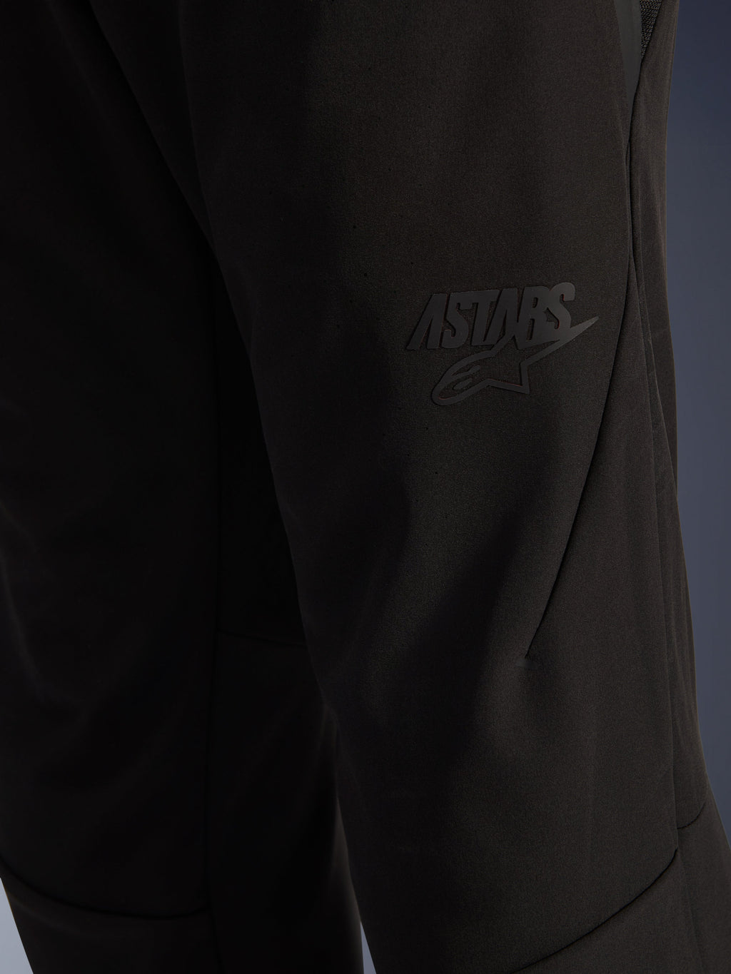 Pantalones Alpinestars Aeroshell Airflow, pantalones de moto negros, primer plano de la zona del muslo con un logotipo ASTARS negro en relieve, material softshell elástico ligero con diseño de costuras ergonómicas