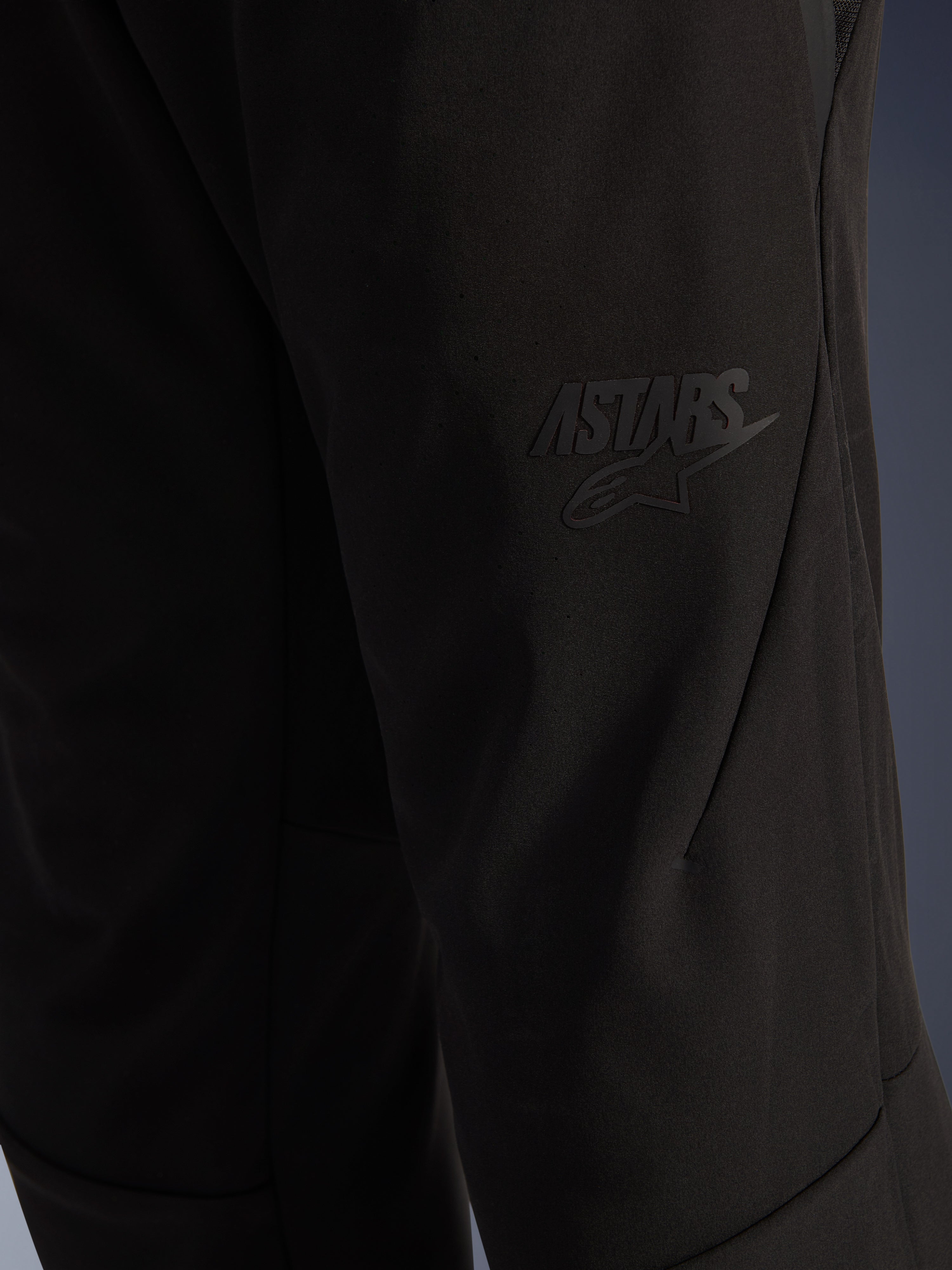 Pantalones Alpinestars Aeroshell Airflow, pantalones de moto negros, primer plano de la zona del muslo con un logotipo ASTARS negro en relieve, material softshell elástico ligero con diseño de costuras ergonómicas