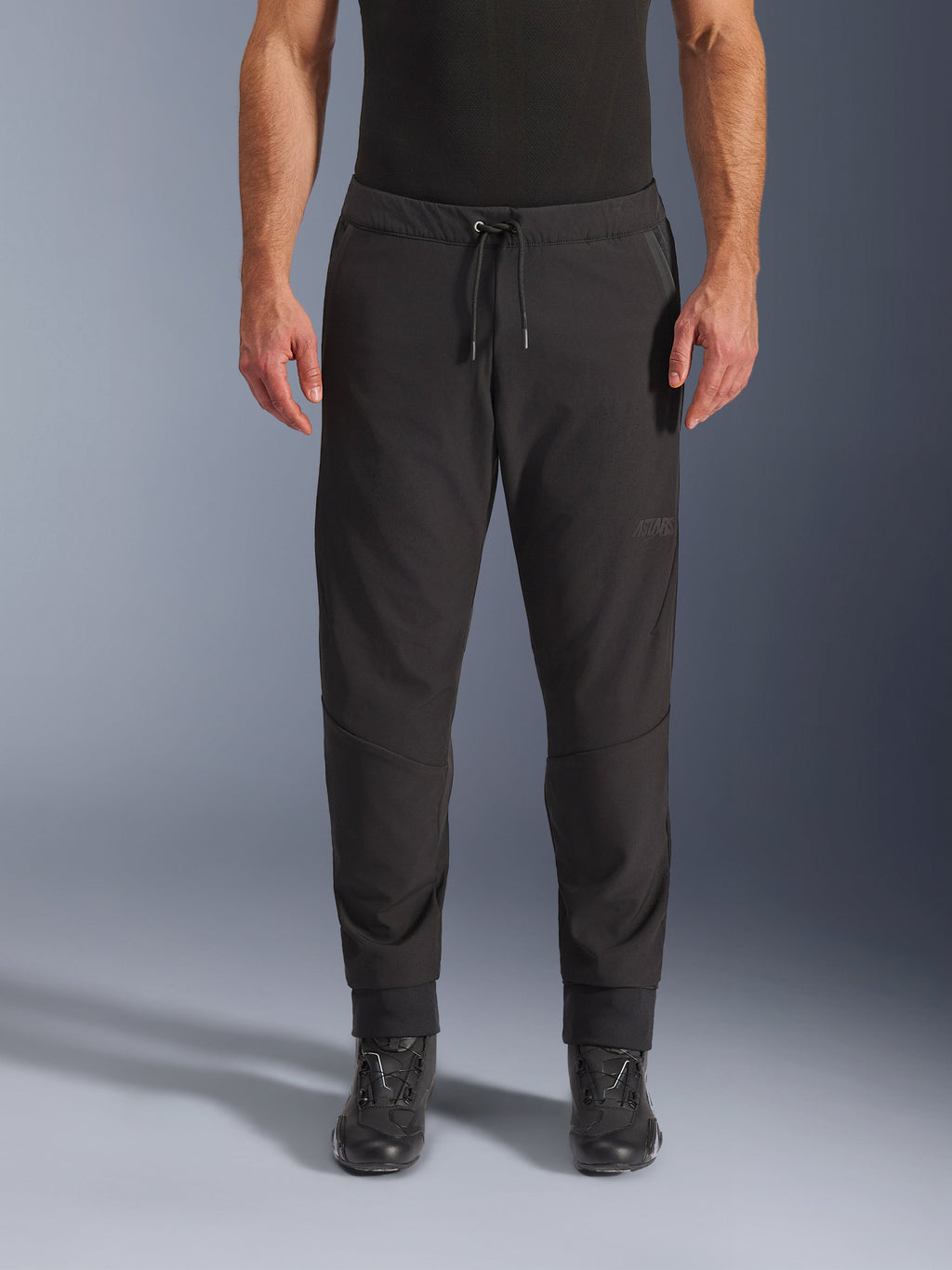 Pantalones Alpinestars Aeroshell Airflow, pantalones de moto negros, vista frontal con botas de conducción negras, cuentan con cintura con cordón y puños de tobillo entallados, diseño de softshell ligero con marca tonal sutil.