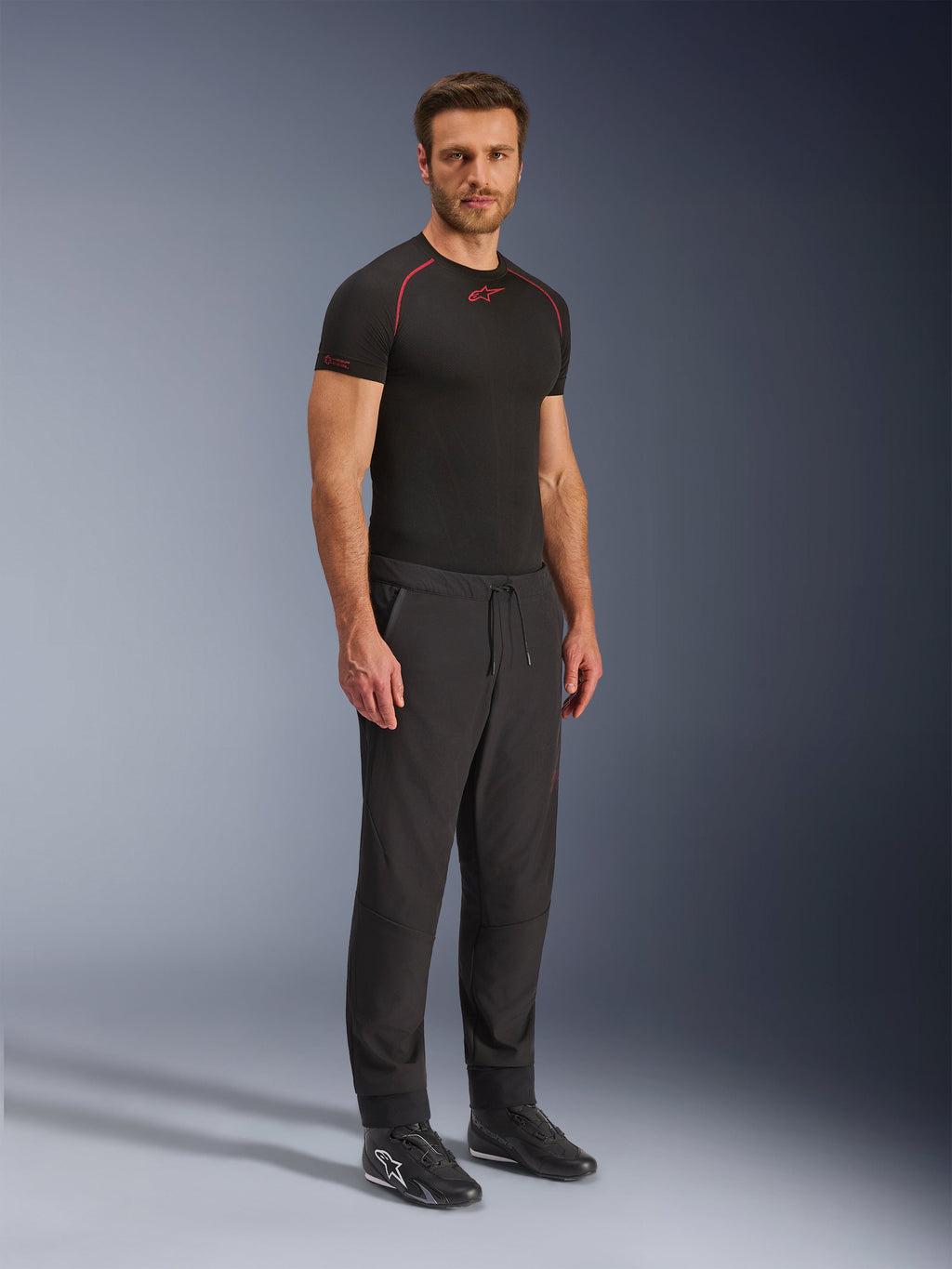 Pantalones Alpinestars Aeroshell Airflow, pantalones de motociclismo negros, toma de estudio de cuerpo completo de un modelo con pantalones softshell de cintura con cordón y puños elásticos, combinados con una camiseta de capa base Alpinestars negra y calzado de conducción negro.