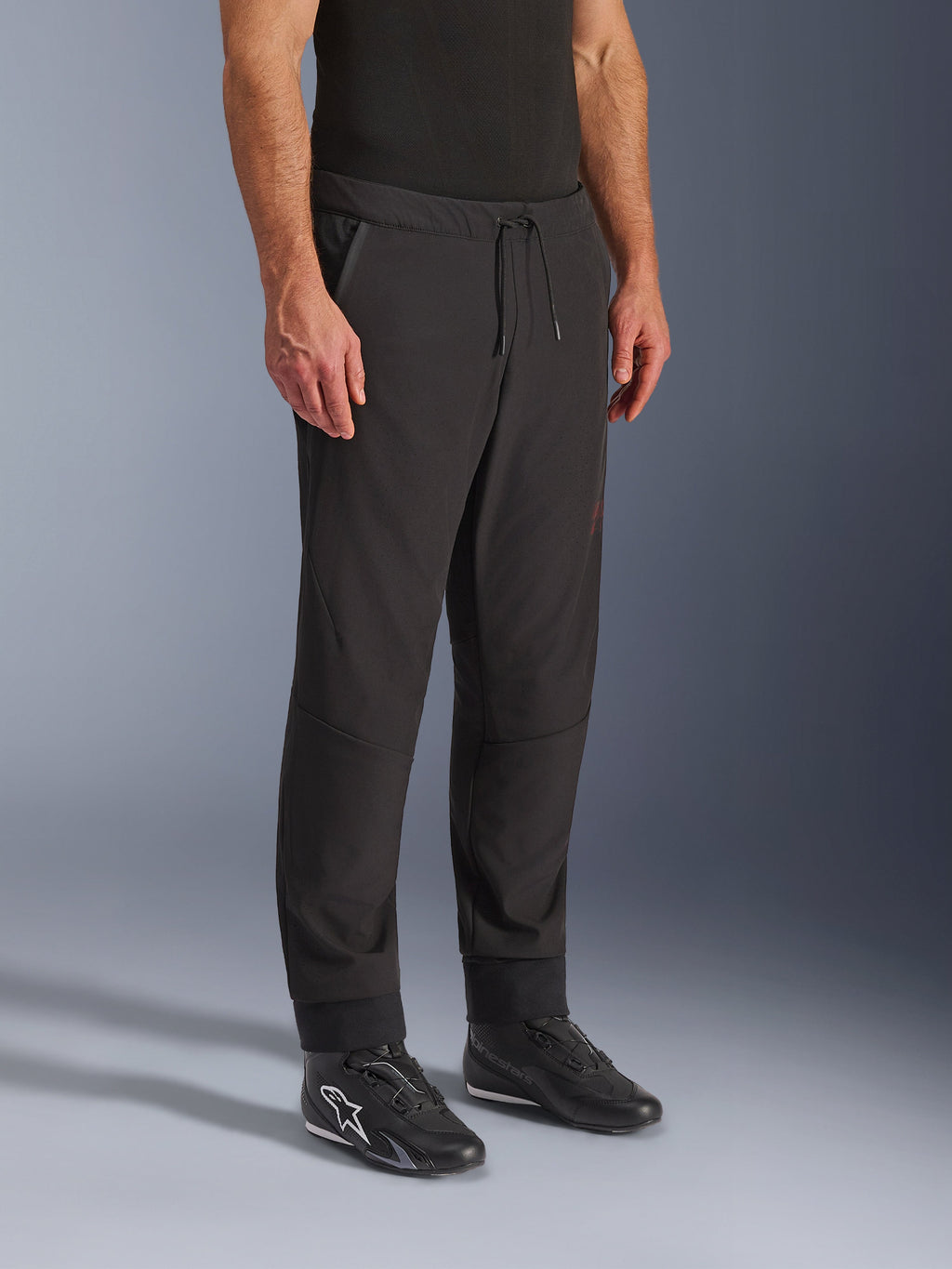 Pantalones Alpinestars Aeroshell Airflow, pantalones de motociclismo negros, usados por un modelo con calzado de conducción negro, con cintura con cordón ajustable, puños en los tobillos y tejido softshell perforado con láser para una máxima ventilación