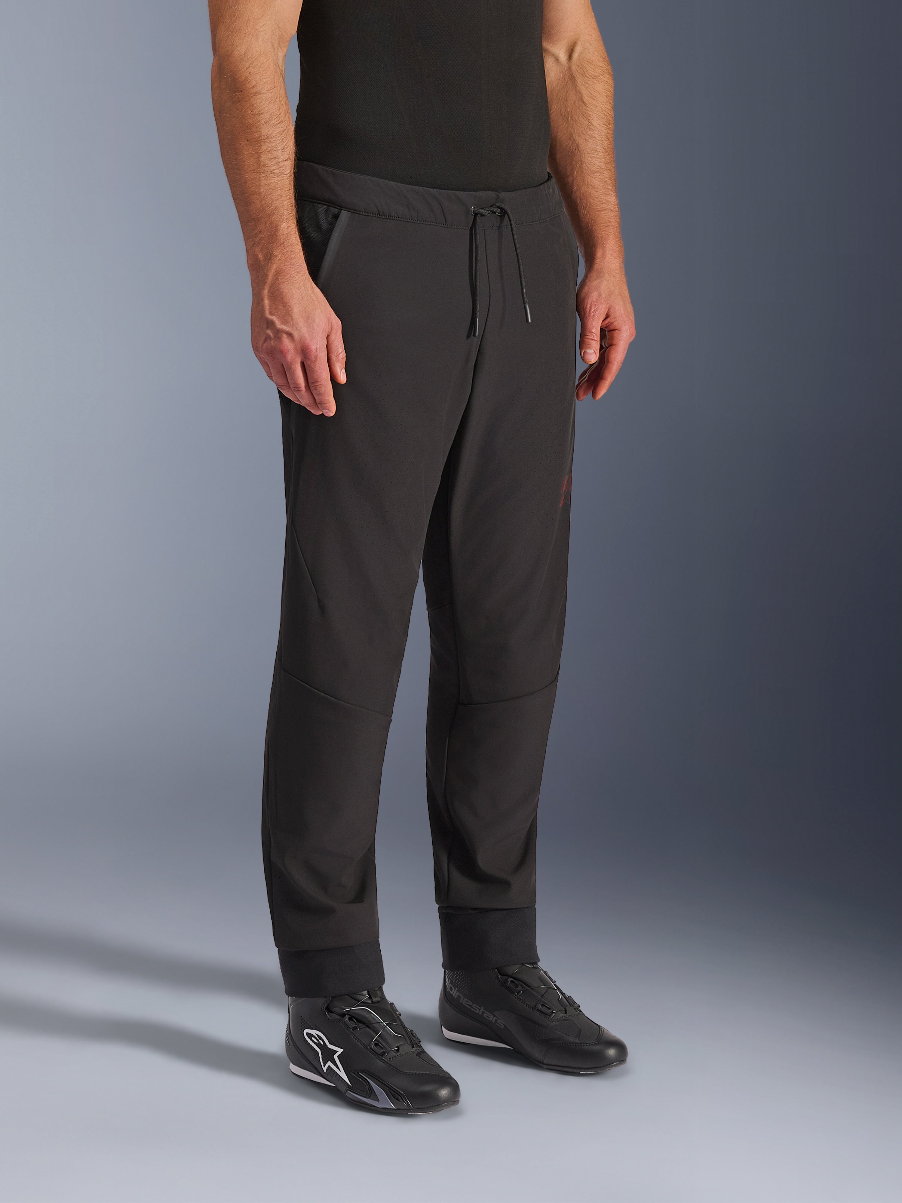 Pantalones Alpinestars Aeroshell Airflow, pantalones de motociclismo negros, usados por un modelo con calzado de conducción negro, con cintura con cordón ajustable, puños en los tobillos y tejido softshell perforado con láser para una máxima ventilación