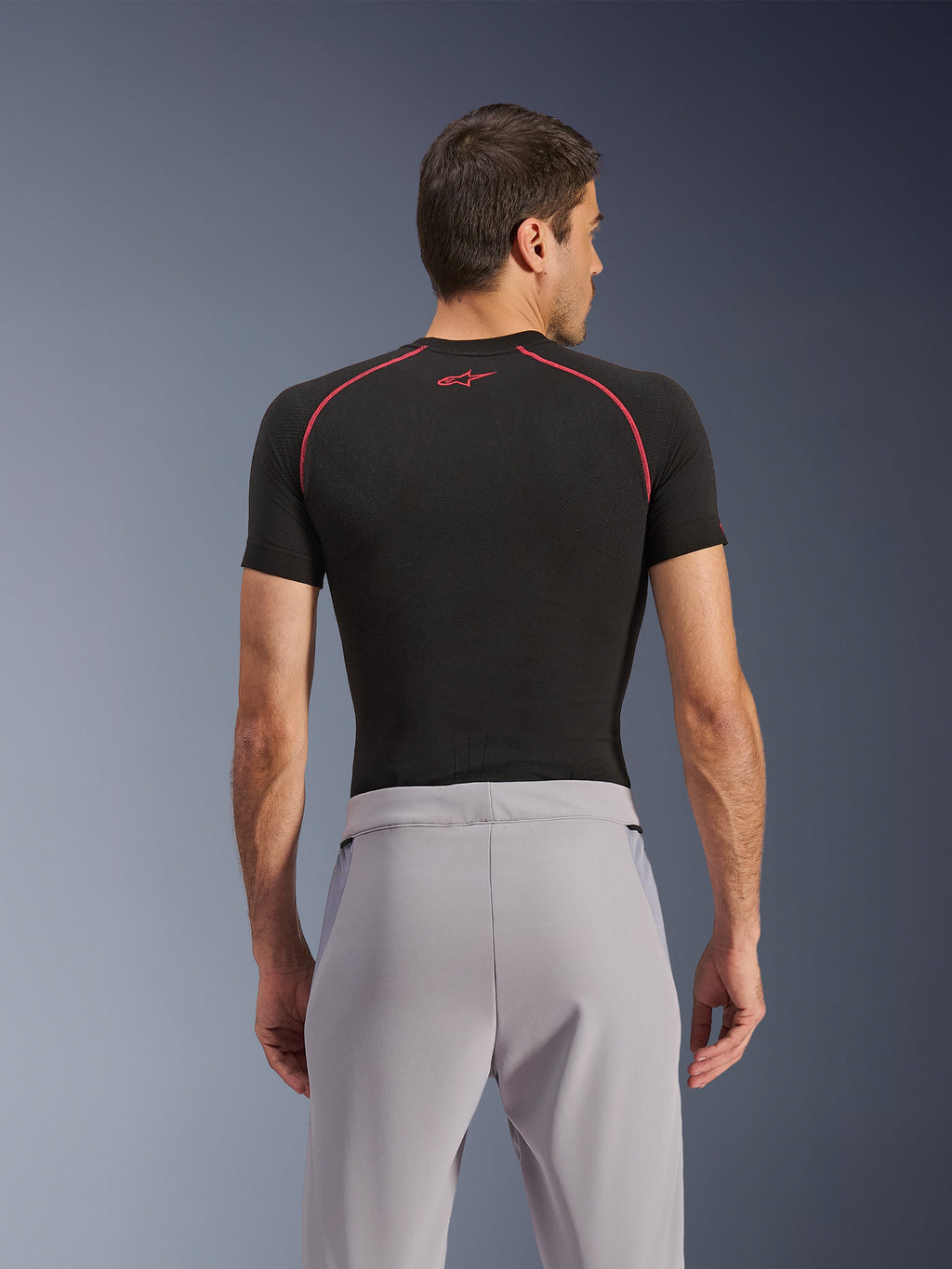 Pantalones Alpinestars Aeroshell Airflow, Gris Medio, vista trasera, combinados con una capa base de manga corta negra con costuras y logotipo rojos, con tejido softshell elástico ligero y paneles de malla para una ventilación máxima, ajuste deportivo ergonómico para la conducción de motocicletas