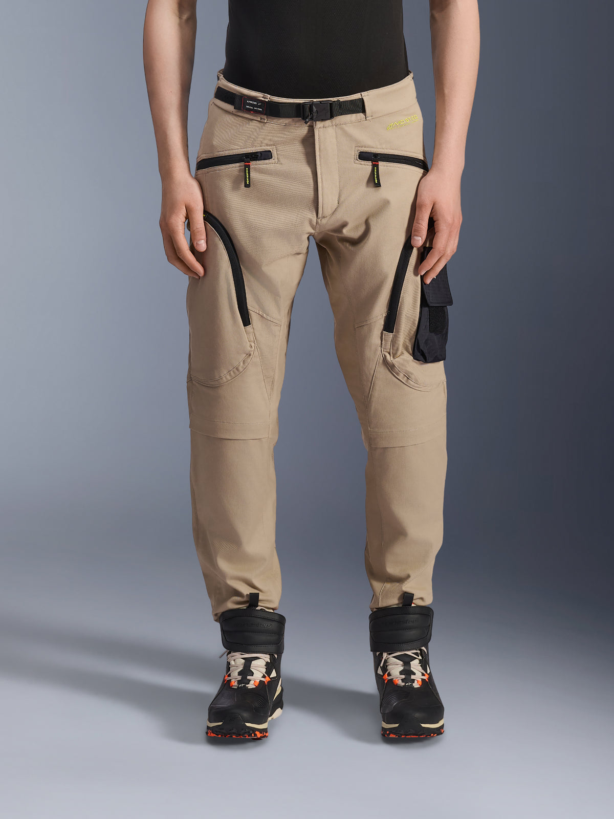 Alpinestars Acteon Pants, pantalones de moto urbanos, color arena, con diseño tipo cargo multibolsillos, cremalleras de espiral inversa negras, cinturón integrado y corte ajustado, combinados con botas negras y naranjas.