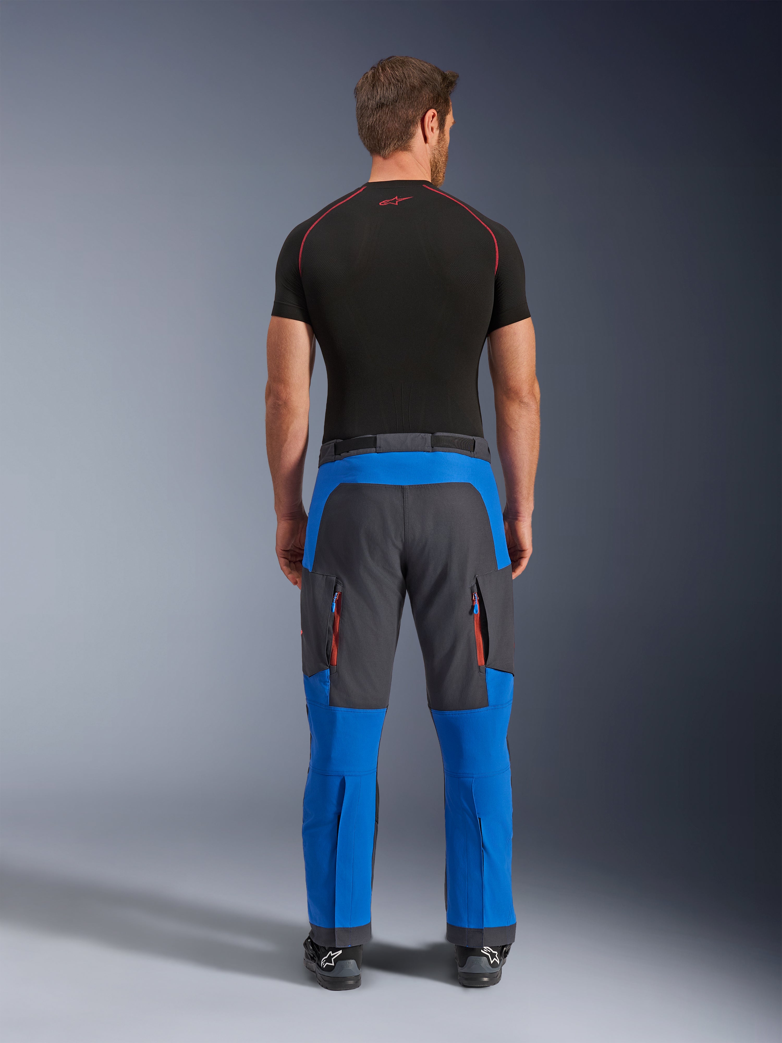 Pantalones Alpinestars Maxdura Dual en azul real y gris oscuro, pantalones de aventura para motociclismo vistos desde atrás, con bolsillos cargo con cremallera y detalles en naranja y diseño sobre la bota, usados con una prenda técnica de manga corta negra