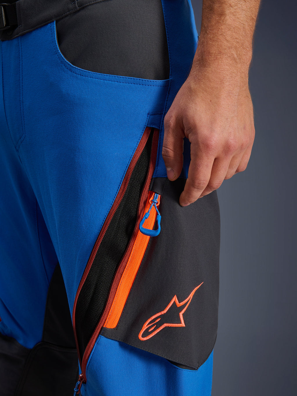 Detalle de los pantalones Alpinestars Maxdura Dual, pantalones de moto de aventura en azul real y gris oscuro, primer plano de la ventilación con cremallera en el muslo con detalles naranjas y tirador azul, logotipo de Alpinestars naranja sobre panel negro, construcción en poliamida elástica resistente