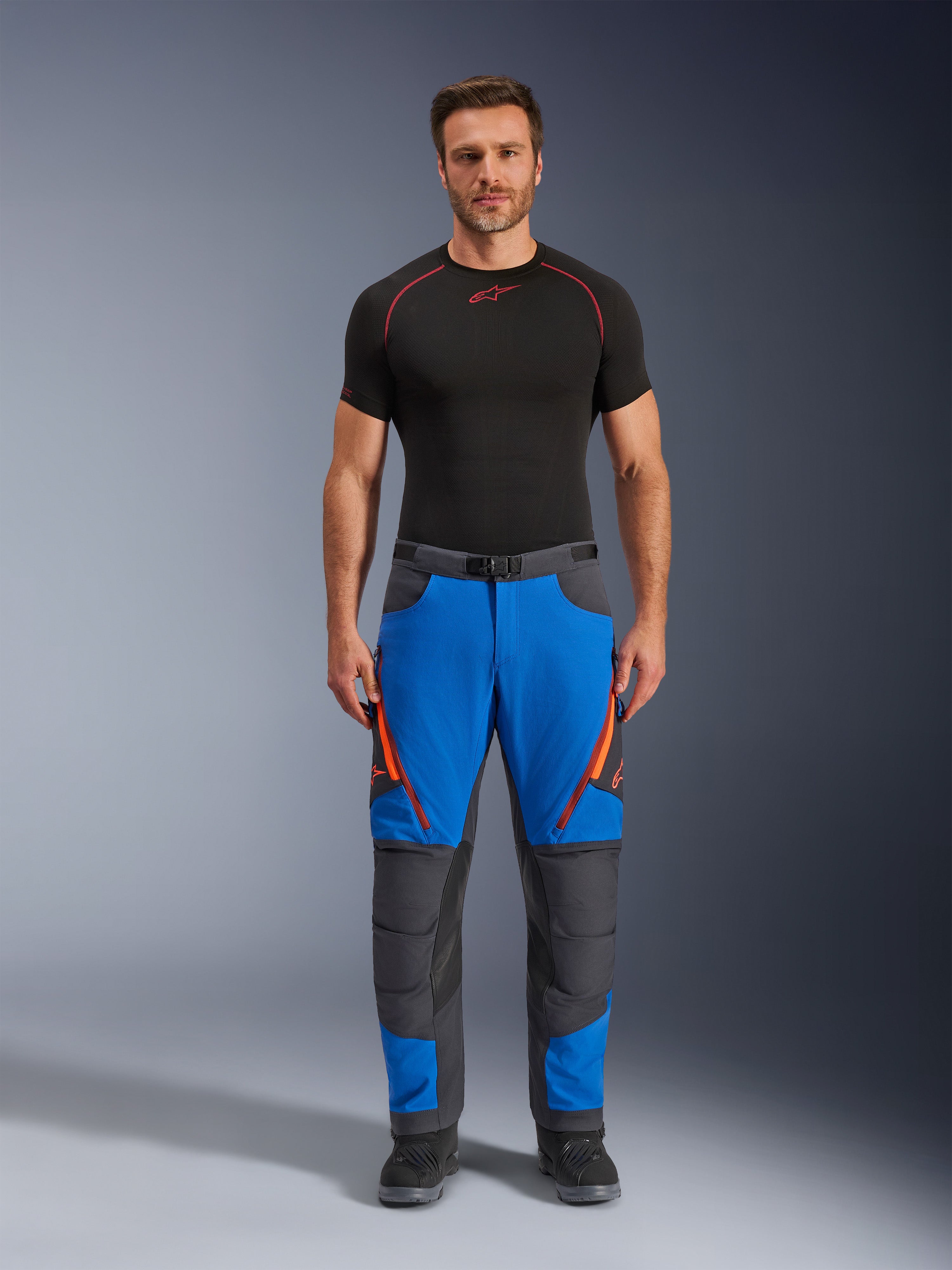 Alpinestars Maxdura Dual Pants, pantalones de moto de aventura, azul real y gris oscuro, detalles de cremalleras naranjas, usados con una capa base Alpinestars negra y botas negras, con paneles reforzados en las rodillas y un diseño técnico off-road