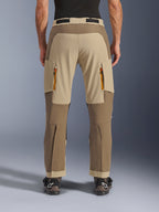 Alpinestars Maxdura Dual Pants, Walnut Aluminum, vista trasera de pantalones de motociclismo de aventura con paneles en tonos tierra y beige, ventilaciones traseras con cremalleras naranjas y diseño sobre la bota, combinados con botas Alpinestars negras.