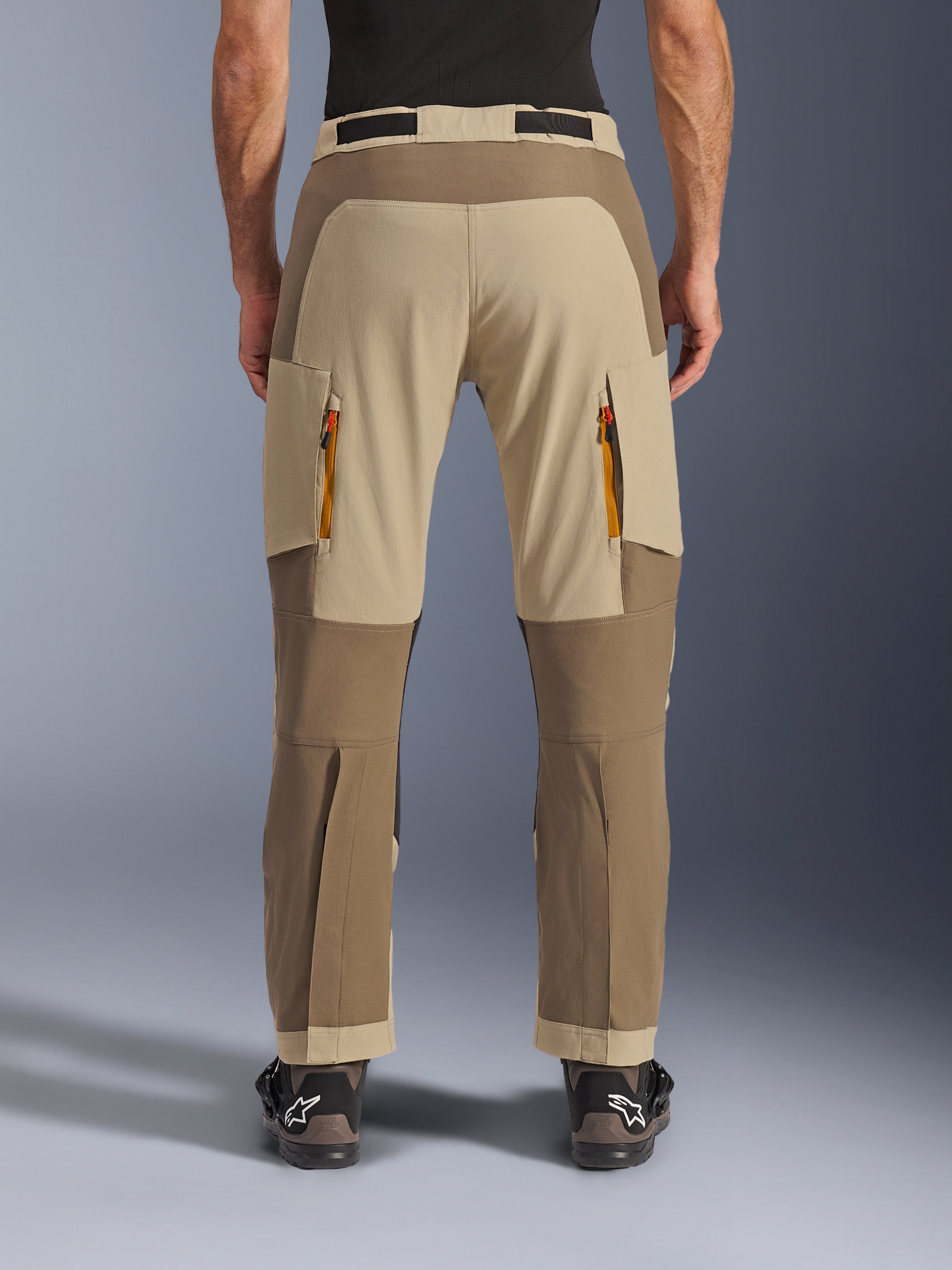 Alpinestars Maxdura Dual Pants, Walnut Aluminum, vista trasera de pantalones de motociclismo de aventura con paneles en tonos tierra y beige, ventilaciones traseras con cremalleras naranjas y diseño sobre la bota, combinados con botas Alpinestars negras.