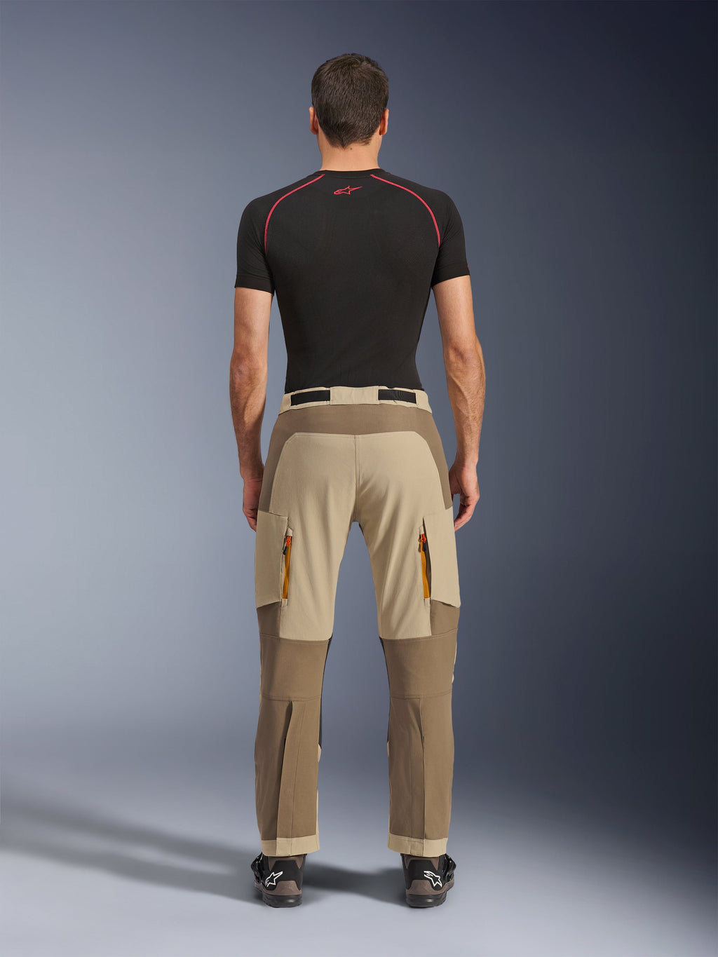 Pantalones Alpinestars Maxdura Dual en Walnut Aluminum, vista trasera que muestra un diseño bitono en marrón y tostado, pantalones para conducción de aventura con bolsillos de carga con cremallera y asiento reforzado, usados con una camiseta de compresión negra y botas de motociclismo.