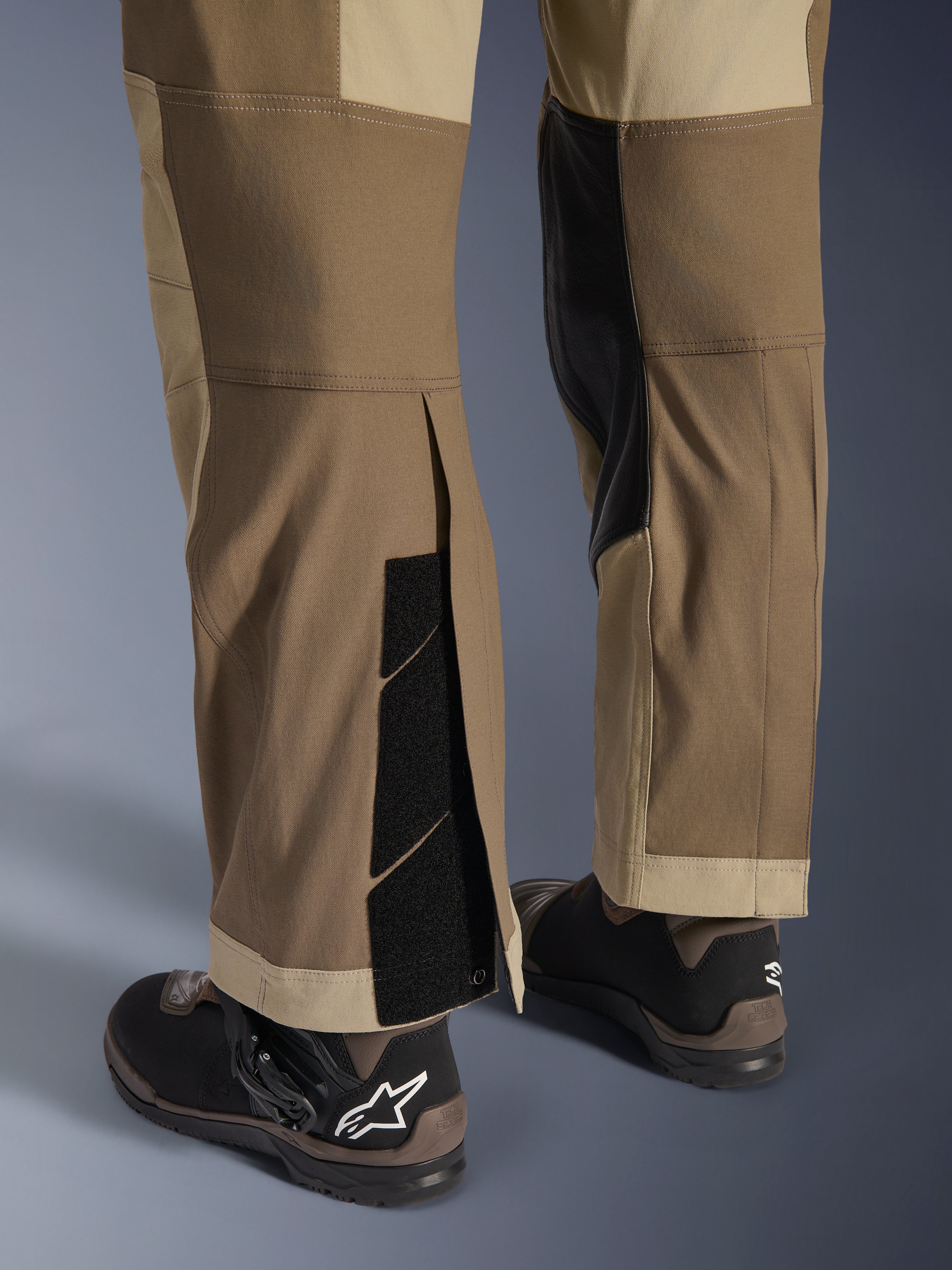 Pantalones Alpinestars Maxdura Dual en Walnut Aluminum, vista detallada de la parte inferior de la pierna y el dobladillo con diseño sobre la bota, cierres de velcro ajustables y paneles interiores reforzados, combinados con botas de moto negras.