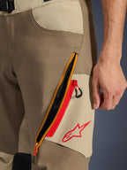 Pantalones Alpinestars Maxdura Dual, Walnut y Aluminum, primer plano del detalle del muslo con un gran sistema de ventilación diagonal con cremallera y detalles rojos, logotipo rojo de Alpinestars sobre un panel tostado, equipamiento de aventura para motocicleta
