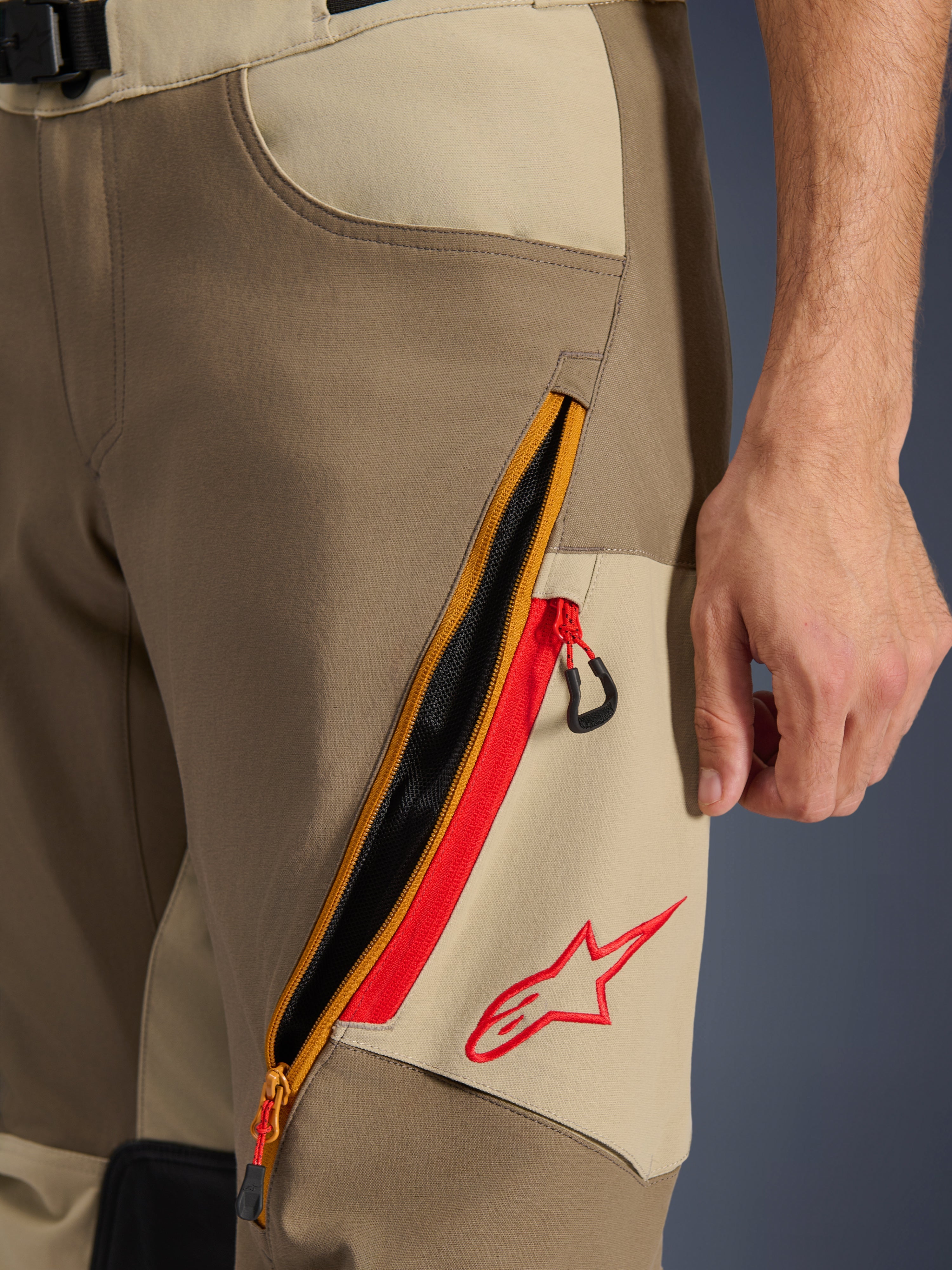 Pantalones Alpinestars Maxdura Dual, Walnut y Aluminum, primer plano del detalle del muslo con un gran sistema de ventilación diagonal con cremallera y detalles rojos, logotipo rojo de Alpinestars sobre un panel tostado, equipamiento de aventura para motocicleta