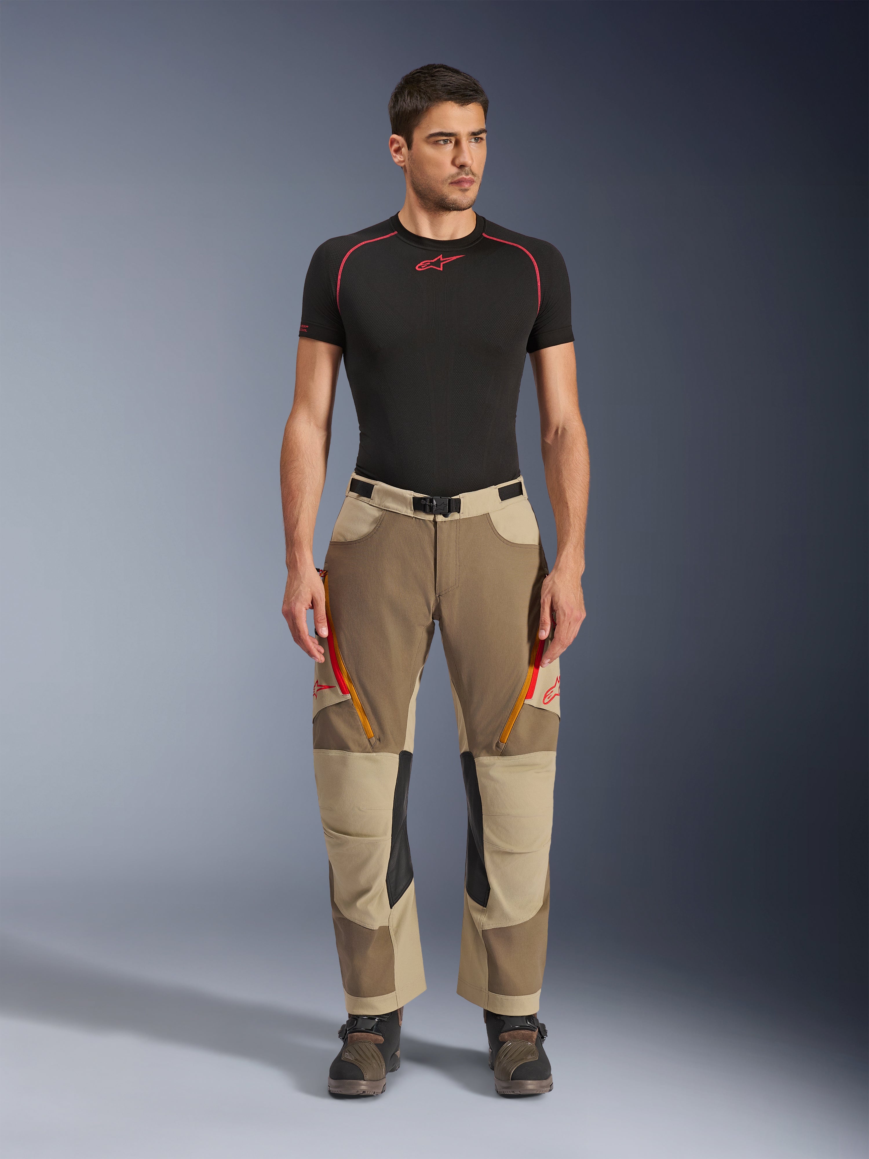 Alpinestars Maxdura Dual Pants, pantalones de conducción Adventure y Dual-Sport, Walnut Aluminum, usados con una camiseta de compresión negra de manga corta y botas de adventure, con ventilaciones con cremallera naranja y logos rojos en los muslos, cinturón negro con hebilla, diseño sobre la bota para conducción de motocicletas.