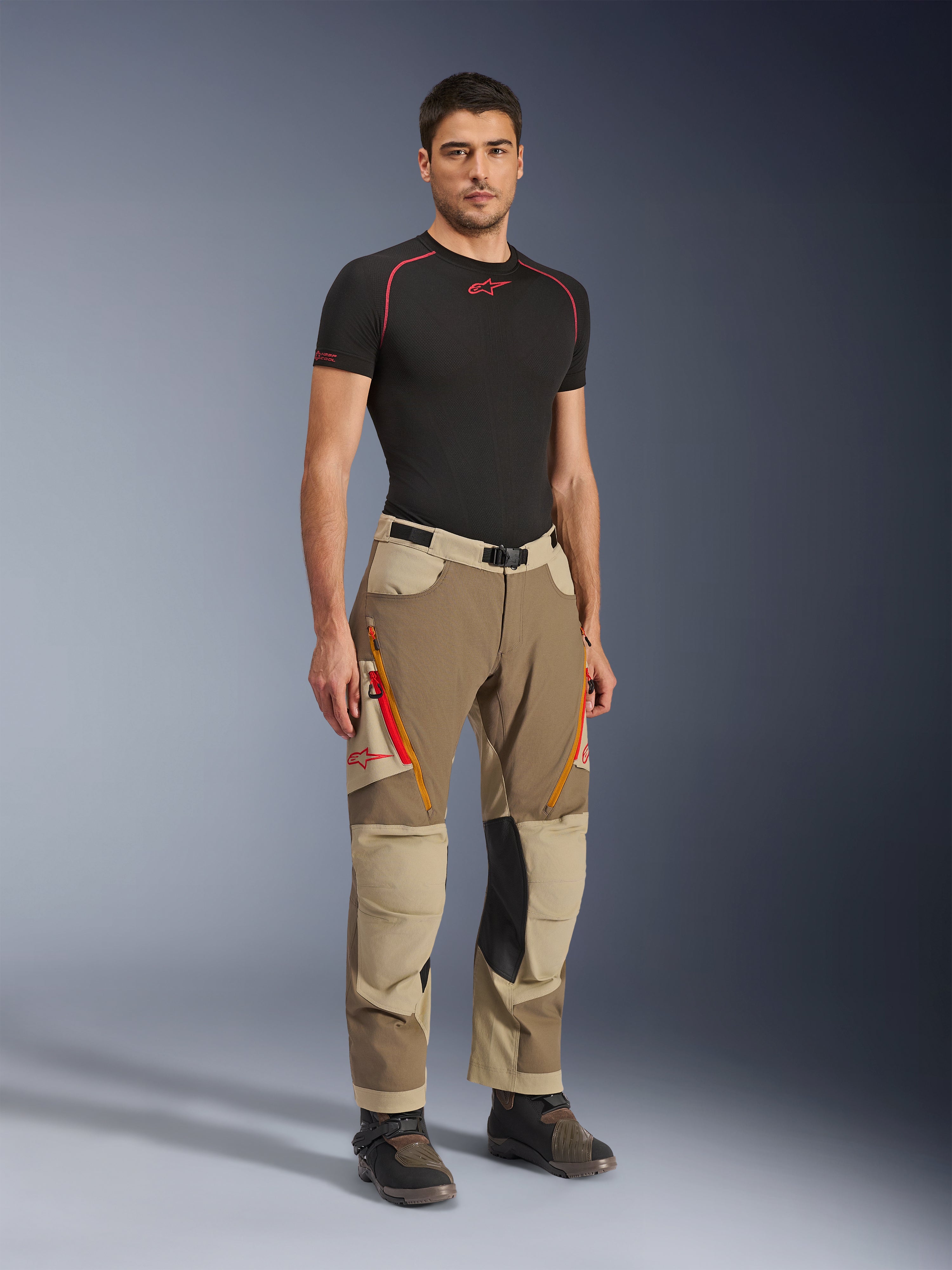 Pantalones Alpinestars Maxdura Dual en Walnut Aluminum, pantalones de moto de aventura en marrón y beige con cremalleras naranjas y paneles de cuero negro en las rodillas, combinados con una camiseta de capa base negra y botas de aventura, vista frontal de cuerpo completo