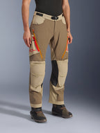 Alpinestars Maxdura Dual Pants, pantalones de aventura en marrón Walnut Aluminum y caqui, con ventilaciones con cremallera en los muslos en naranja y rojo, paneles interiores de las rodillas en cuero negro reforzado y logotipo Astars rojo, usados con botas técnicas.