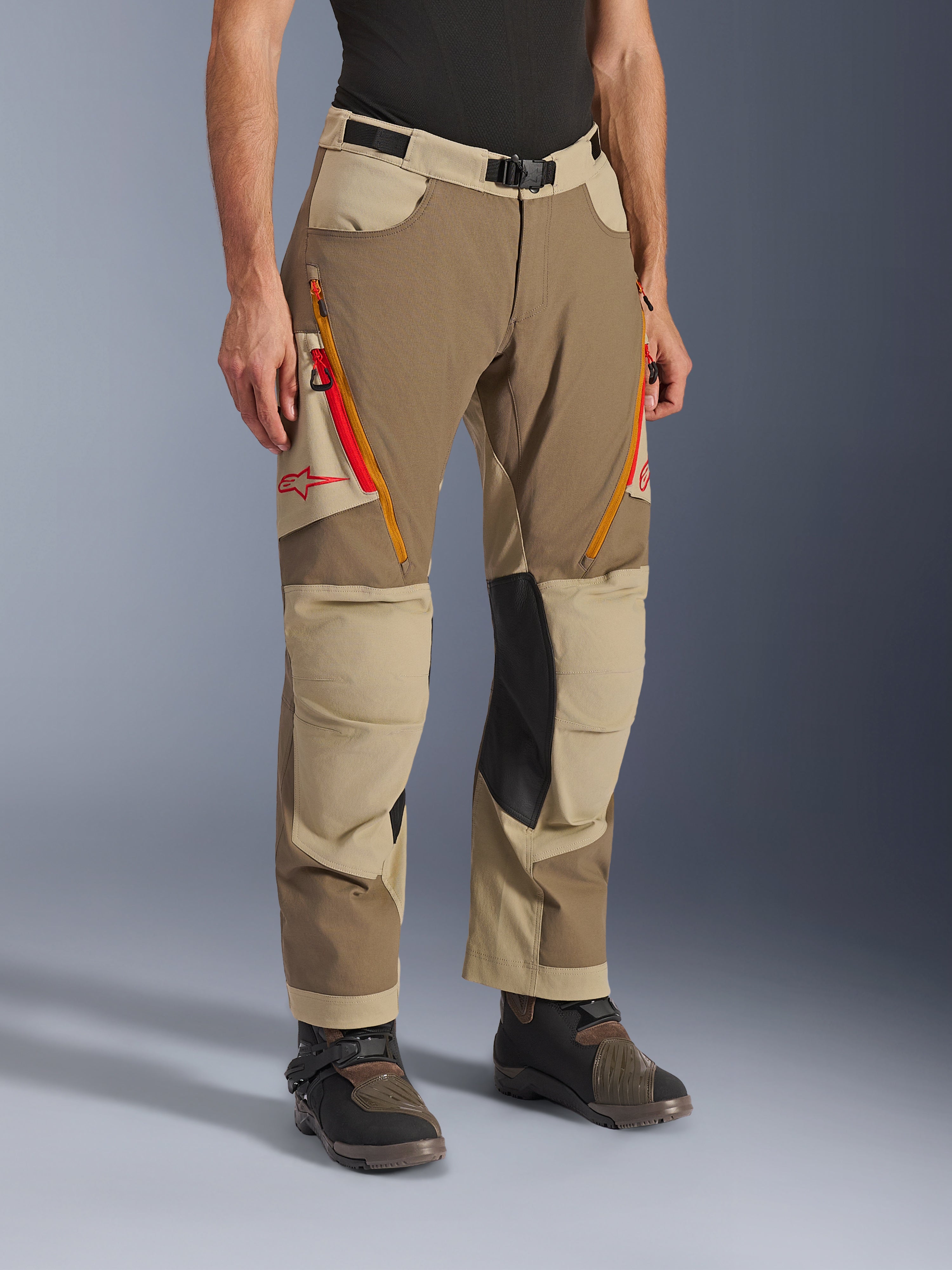 Alpinestars Maxdura Dual Pants, pantalones de aventura en marrón Walnut Aluminum y caqui, con ventilaciones con cremallera en los muslos en naranja y rojo, paneles interiores de las rodillas en cuero negro reforzado y logotipo Astars rojo, usados con botas técnicas.