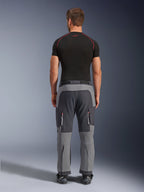 Alpinestars Maxdura Dual Pants, pantalones de motociclismo de aventura en gris oscuro y gris profundo, vista trasera en modelo con camiseta negra y botas, con paneles grises multitono y bolsillos de carga con cremallera y detalles rojos.