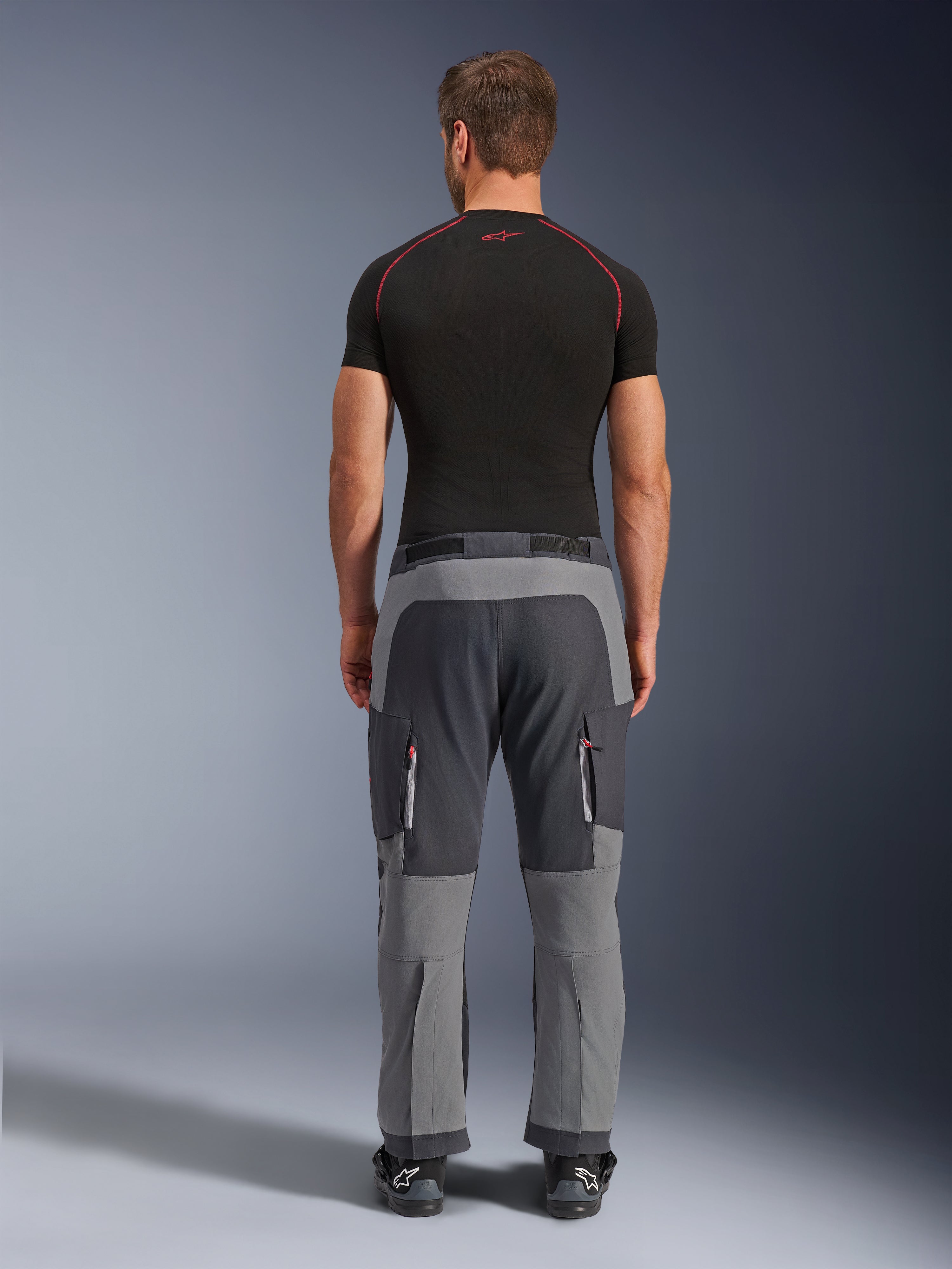 Alpinestars Maxdura Dual Pants, pantalones de motociclismo de aventura en gris oscuro y gris profundo, vista trasera en modelo con camiseta negra y botas, con paneles grises multitono y bolsillos de carga con cremallera y detalles rojos.