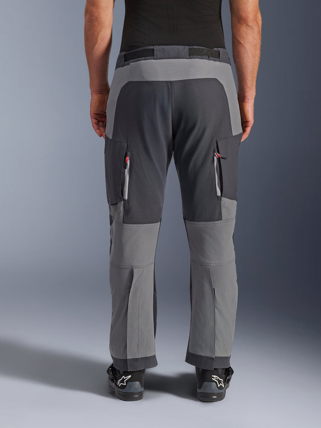 Pantalones Alpinestars Maxdura Dual, pantalones de aventura para moto, gris oscuro y gris intenso, vista trasera que resalta los paneles reforzados en el asiento y las rodillas, ventilaciones con cremallera en los muslos, usados con botas Alpinestars negras, construcción de poliamida elástica de alta resistencia para conducción dual-sport.