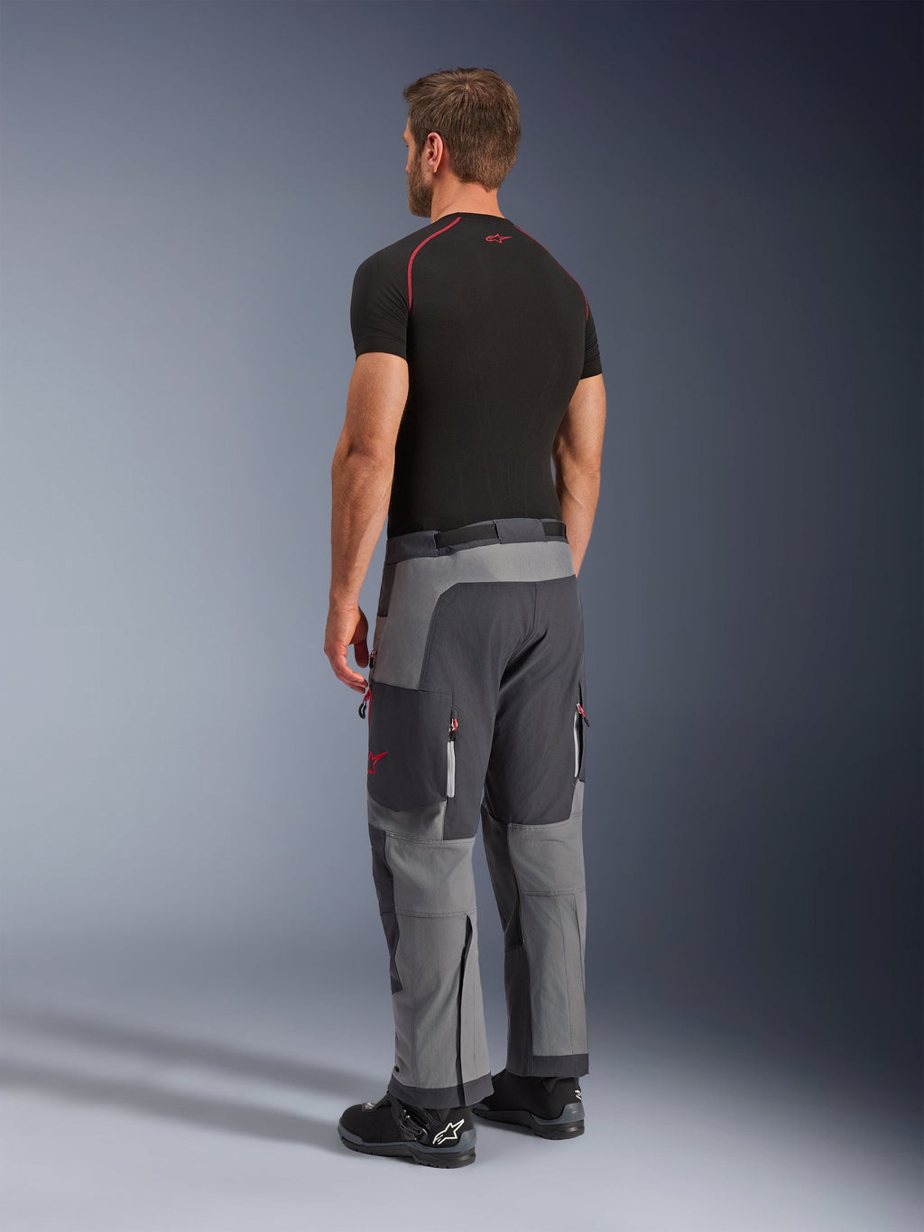 Pantalones Alpinestars Maxdura Dual, pantalones de aventura para motociclismo, gris oscuro y gris intenso, usados por un modelo en una vista lateral trasera, con detalles rojos en las cremalleras y el logotipo, paneles grises reforzados, bolsillos tipo cargo y ajuste sobre la bota