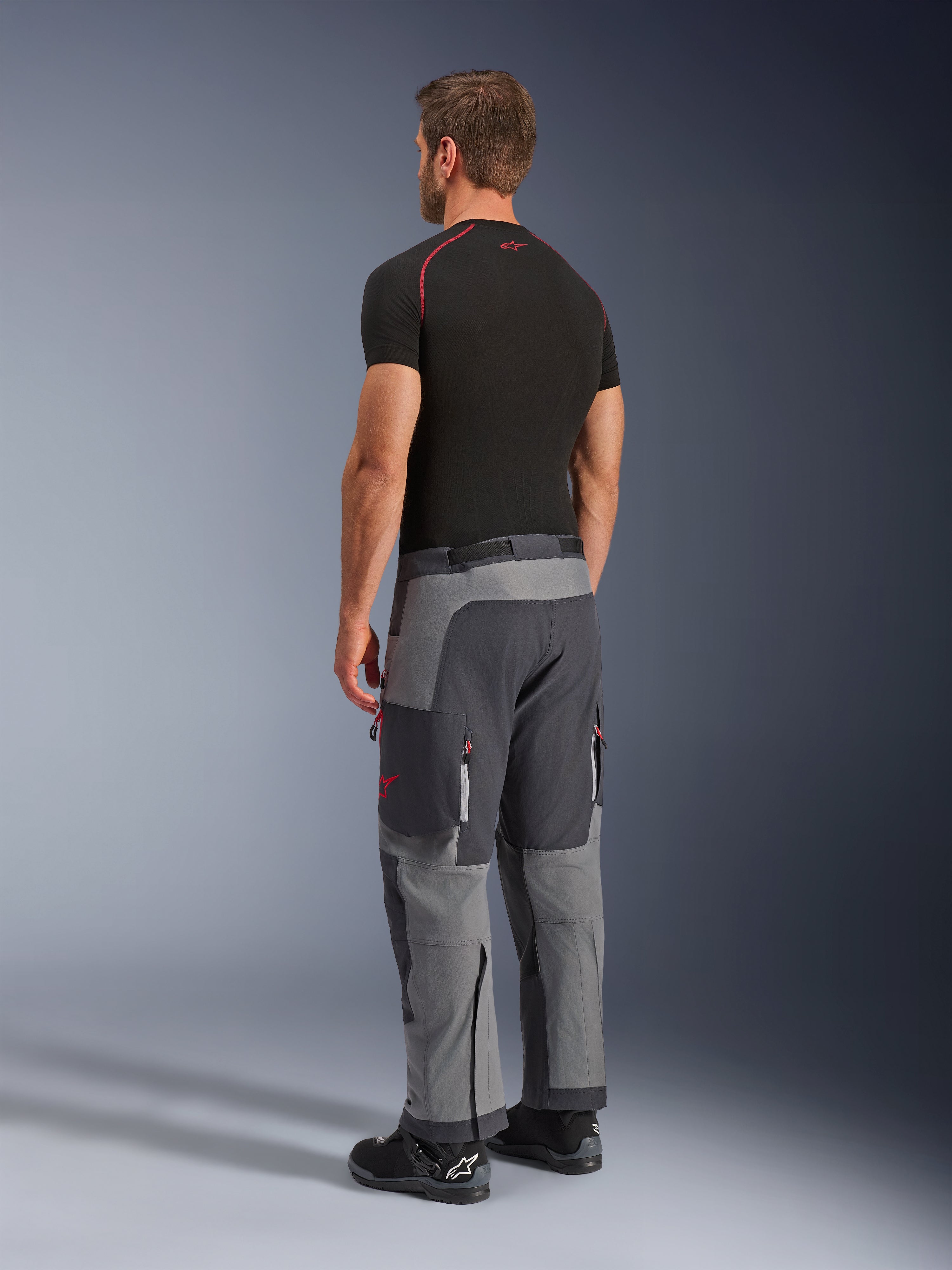 Pantalones Alpinestars Maxdura Dual, pantalones de aventura para motociclismo, gris oscuro y gris intenso, usados por un modelo en una vista lateral trasera, con detalles rojos en las cremalleras y el logotipo, paneles grises reforzados, bolsillos tipo cargo y ajuste sobre la bota