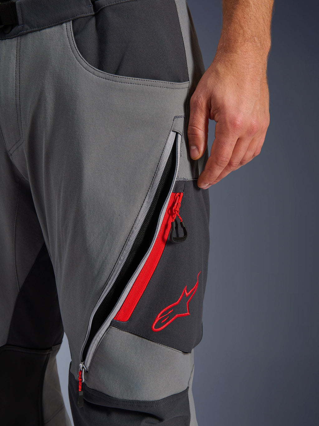 Pantalones Alpinestars Maxdura Dual, pantalones de motociclismo de aventura en gris oscuro y gris intenso, vista de cerca de la ventilación del muslo abierta con cremallera, detalles en rojo y logotipo de estrella roja bordado, diseño de tejido elástico técnico