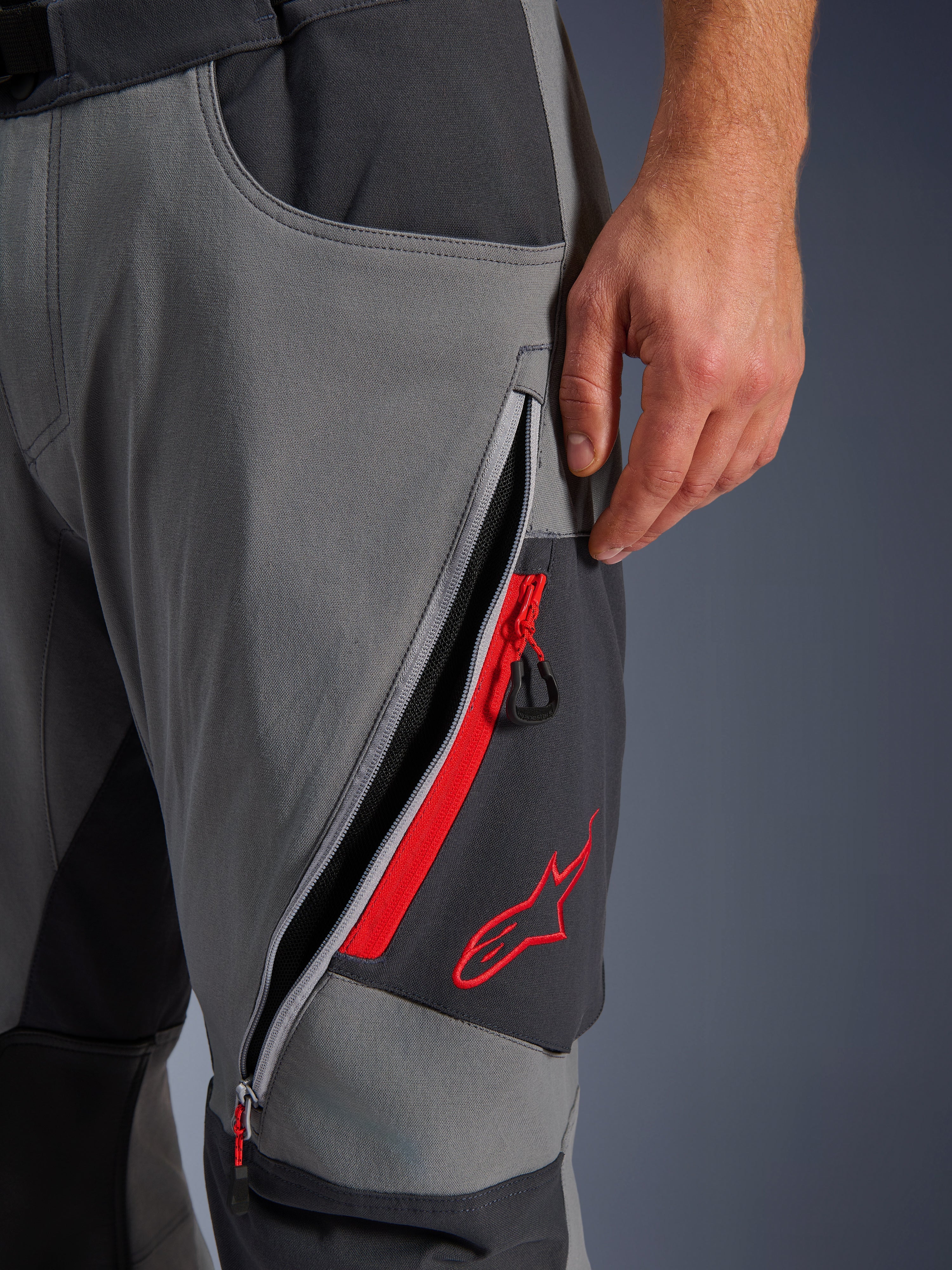 Pantalones Alpinestars Maxdura Dual, pantalones de motociclismo de aventura en gris oscuro y gris intenso, vista de cerca de la ventilación del muslo abierta con cremallera, detalles en rojo y logotipo de estrella roja bordado, diseño de tejido elástico técnico