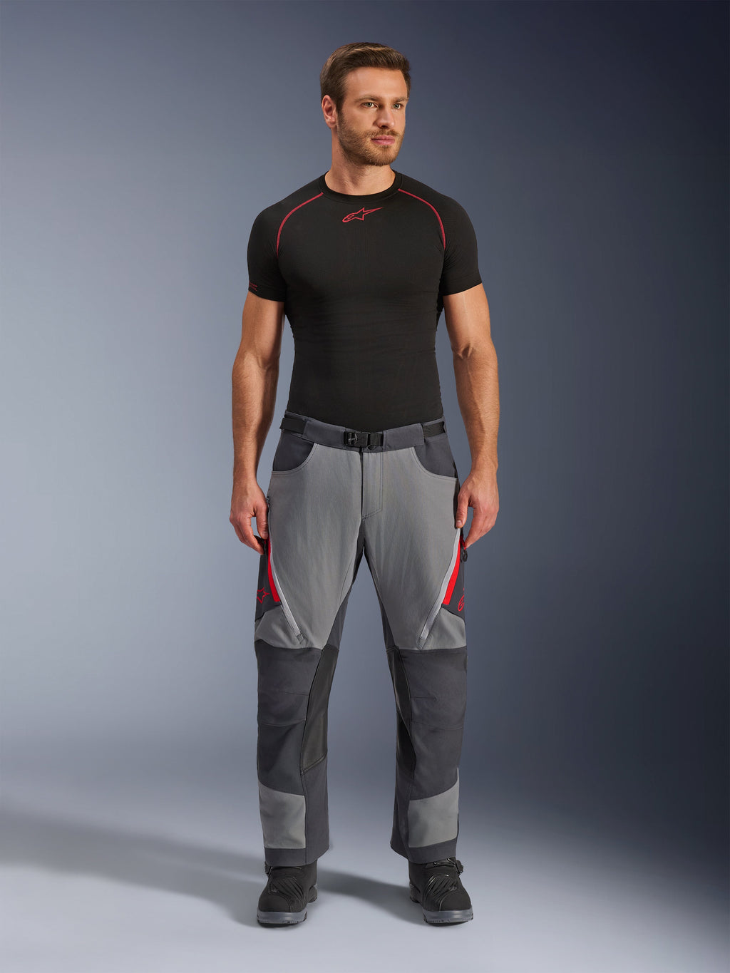 Pantalones Alpinestars Maxdura Dual, pantalones de motociclista, gris oscuro y gris profundo con detalles en rojo, vista frontal usada por un modelo con una capa base Alpinestars negra y botas negras, con paneles reforzados y ventilaciones con cremallera en los muslos, equipamiento de conducción estilo aventura