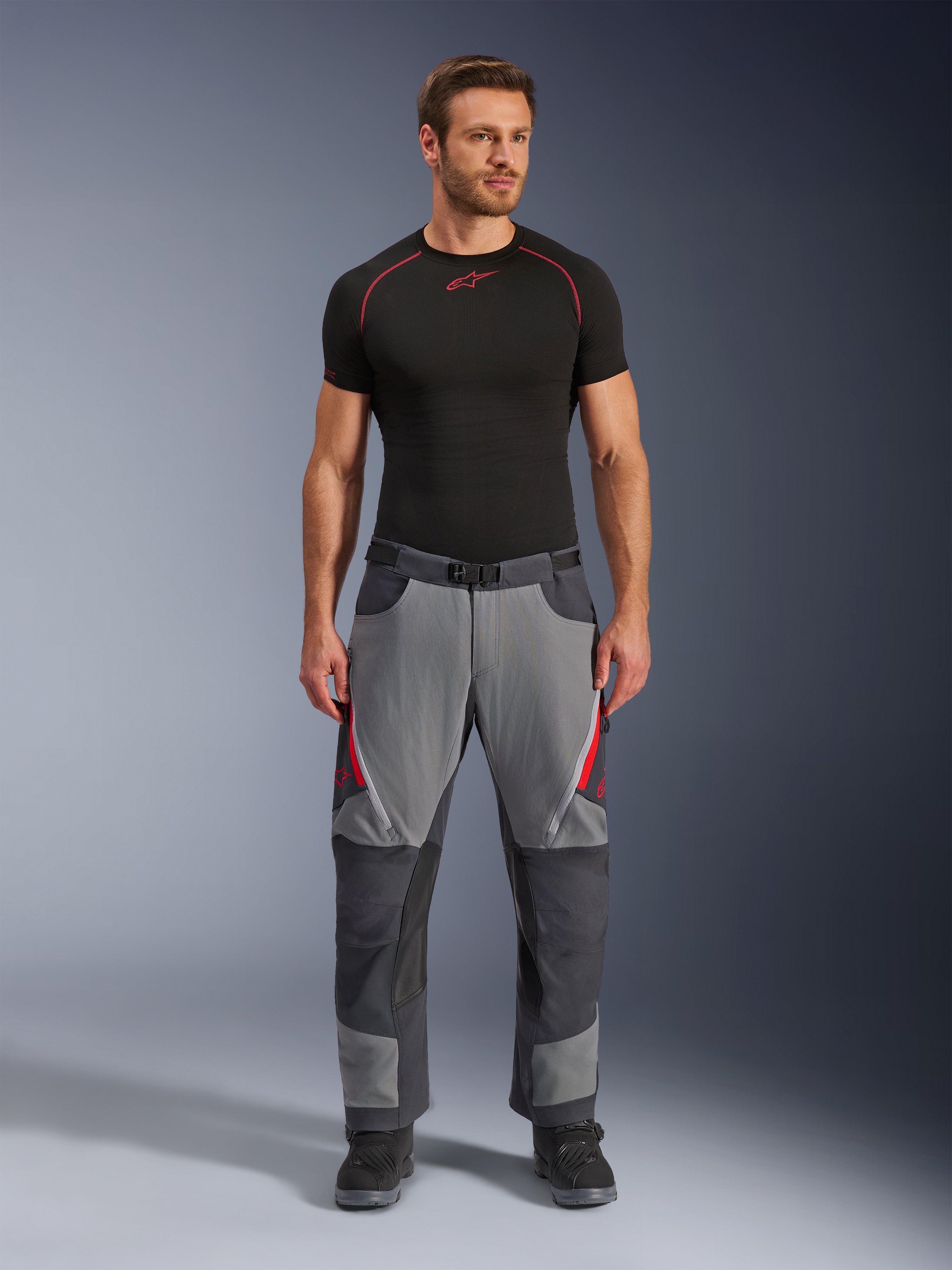 Pantalones Alpinestars Maxdura Dual, pantalones de motociclista, gris oscuro y gris profundo con detalles en rojo, vista frontal usada por un modelo con una capa base Alpinestars negra y botas negras, con paneles reforzados y ventilaciones con cremallera en los muslos, equipamiento de conducción estilo aventura