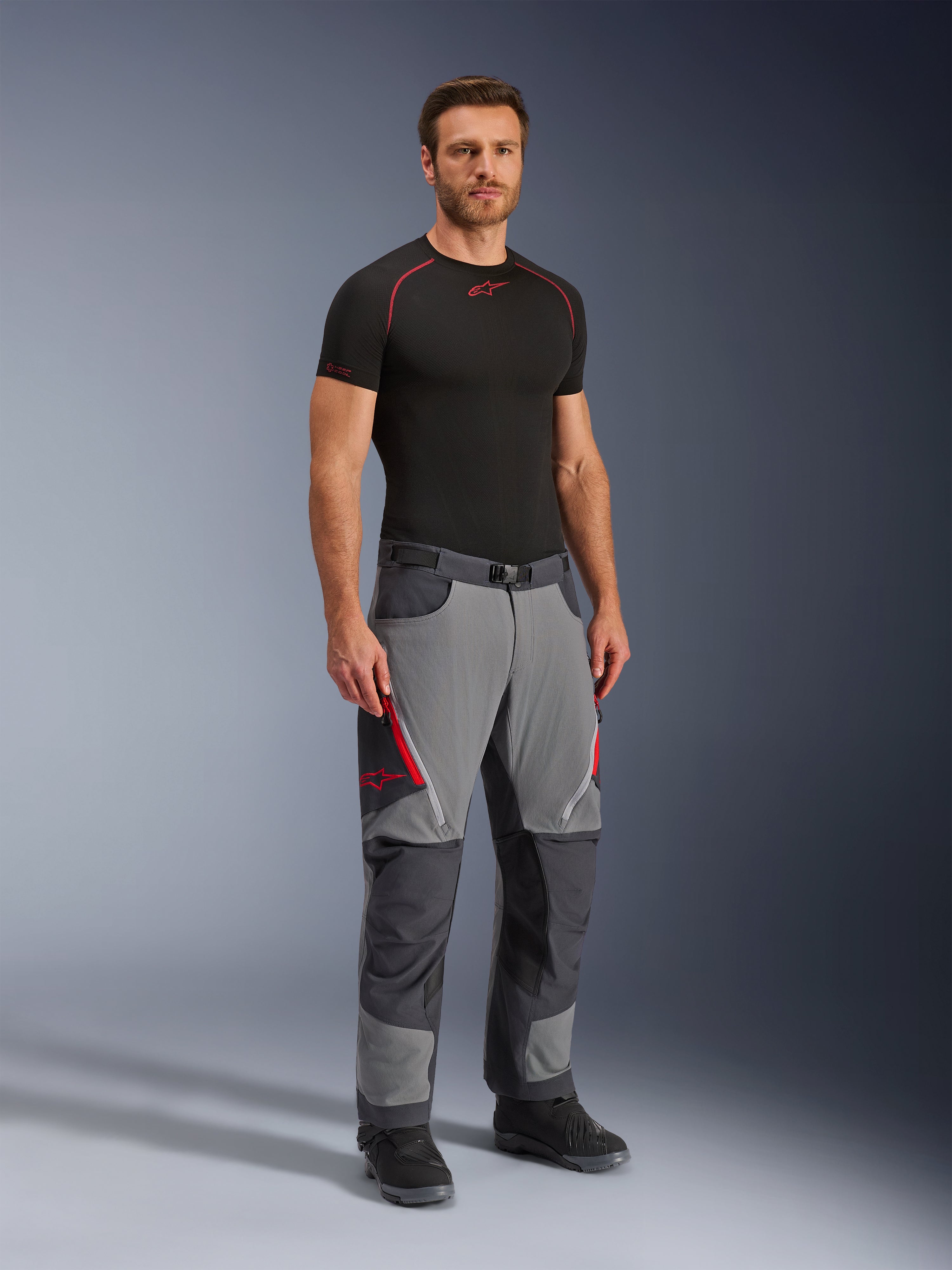 Pantalones Alpinestars Maxdura Dual, pantalones de moto de aventura, gris oscuro y gris intenso, usados con una capa base negra y botas, con aberturas de ventilación rojas con cremallera, paneles reforzados en las rodillas y el asiento, y cintura con cinturón, ajuste atlético para conducción dual-sport