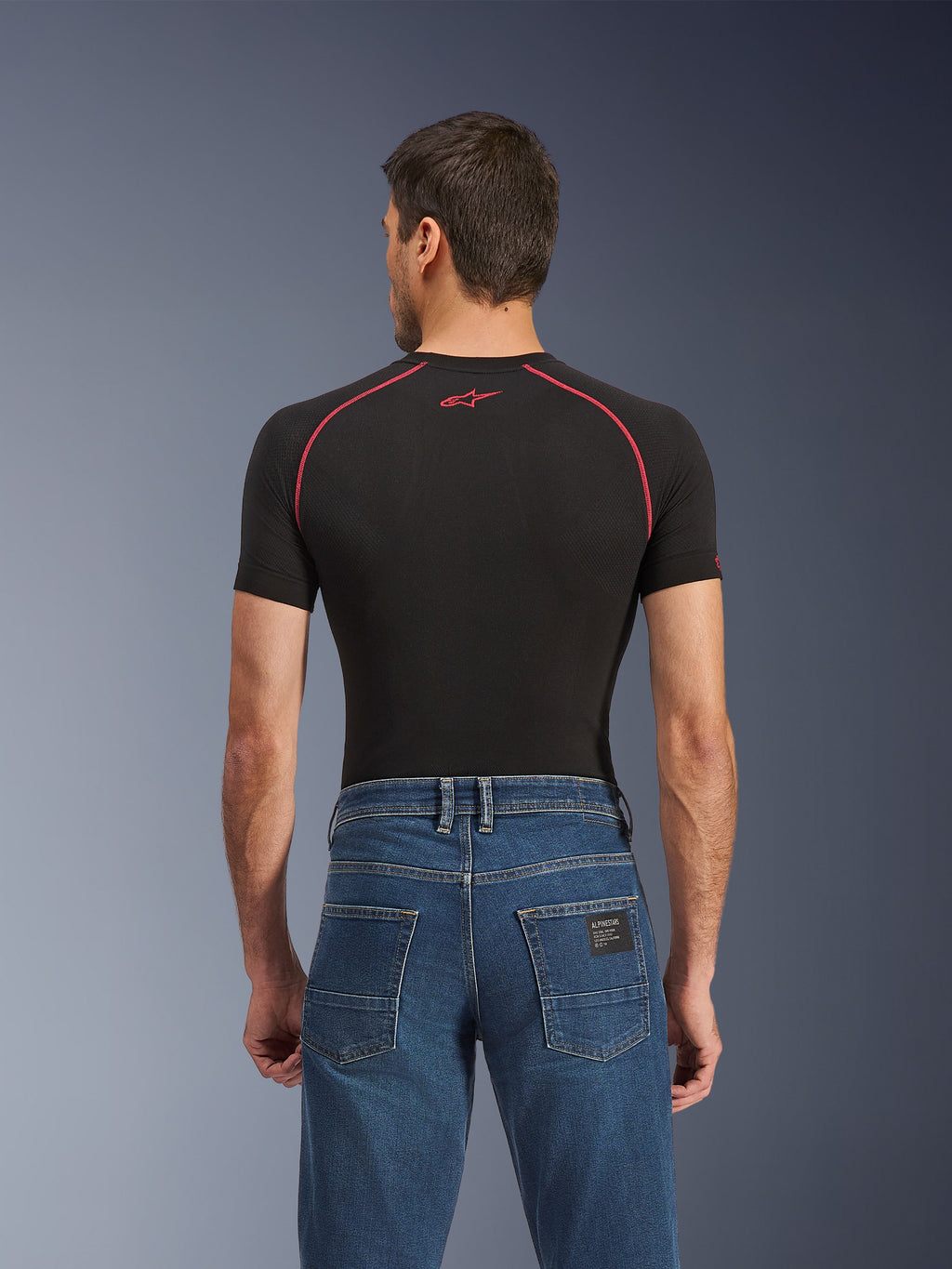 Alpinestars Flex-AST 5 Denim Pants - Short, vaqueros de moto azul lavado índigo, vista trasera con bolsillos posteriores y una pequeña etiqueta negra de Alpinestars, usados con una camiseta de compresión negra con costuras rojas, equipamiento urbano casual para montar en moto.