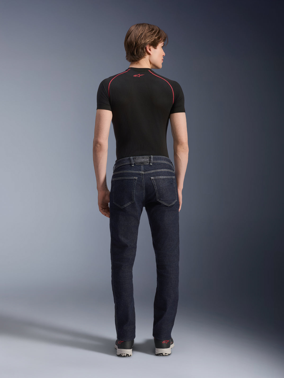 Pantalones Alpinestars Copper V3 Denim en color Rinse Blue, vaqueros de motociclismo urbanos vistos desde atrás, con un ajuste regular de pernera recta y bolsillos traseros, usados con una camiseta negra de manga corta y zapatillas.