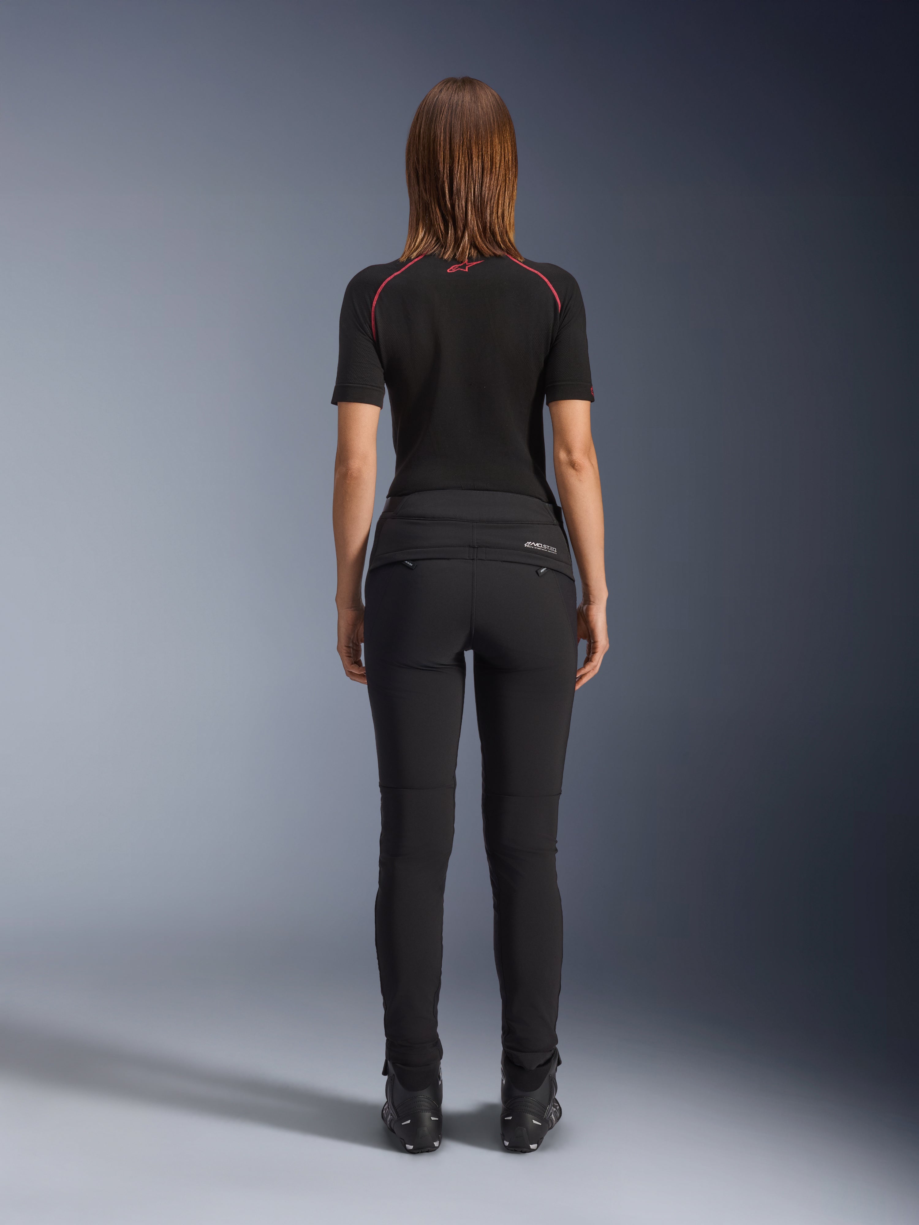 Pantalones Alpinestars Alamosa para mujer, negros, vista trasera que muestra su confección textil de ajuste ceñido, bolsillos traseros con cremallera y marca reflectante MO.ST.EQ. en la pieza trasera del yugo, usados con una prenda base de manga corta negra y botas de moto negras