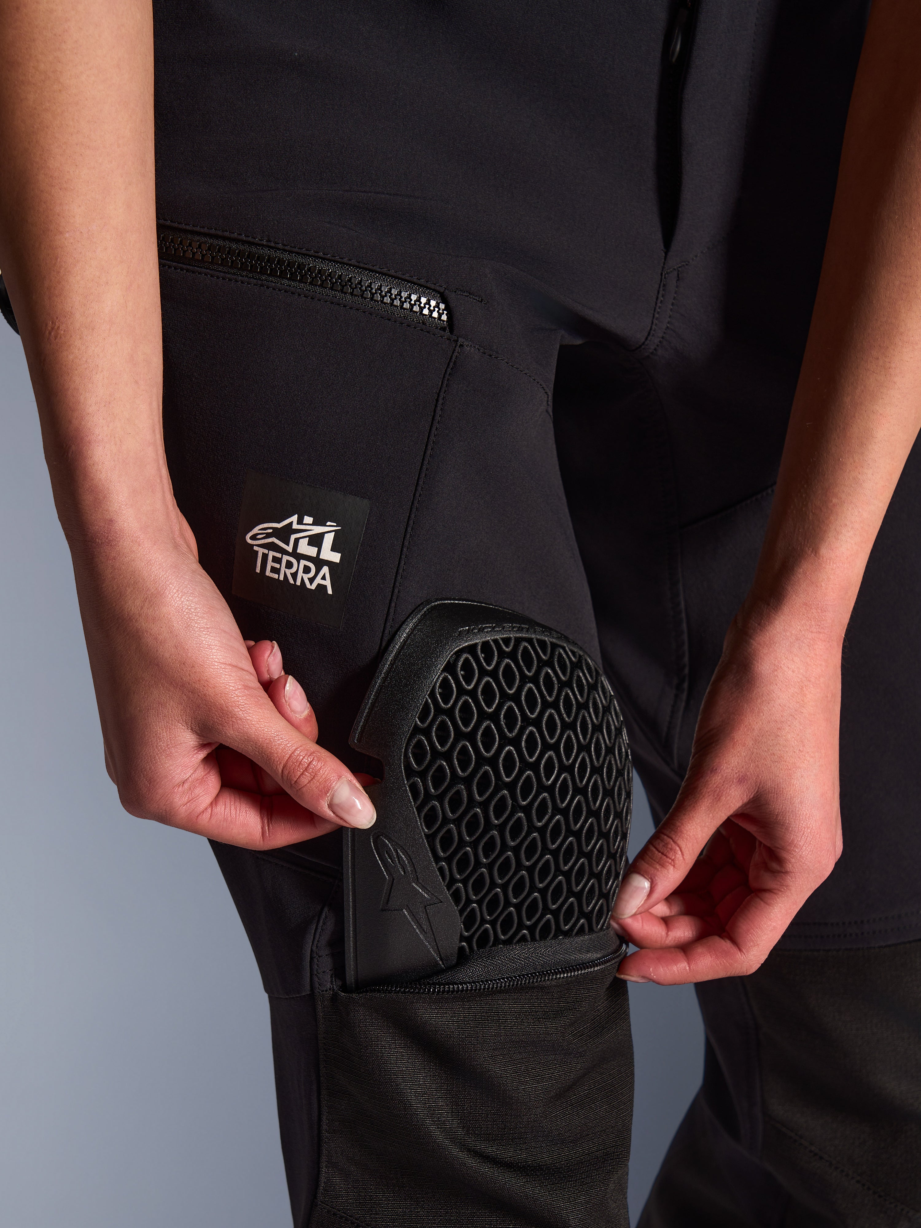Primer plano de los pantalones Alpinestars Stella Flex-AST Explorer en negro, mostrando el uso del bolsillo externo para protectores al insertar la armadura de rodilla Nucleon Flex Plus, detalle de la protección con patrón de panal, logotipo 'ALL TERRA' en el muslo, equipamiento textil funcional de aventura para mujer