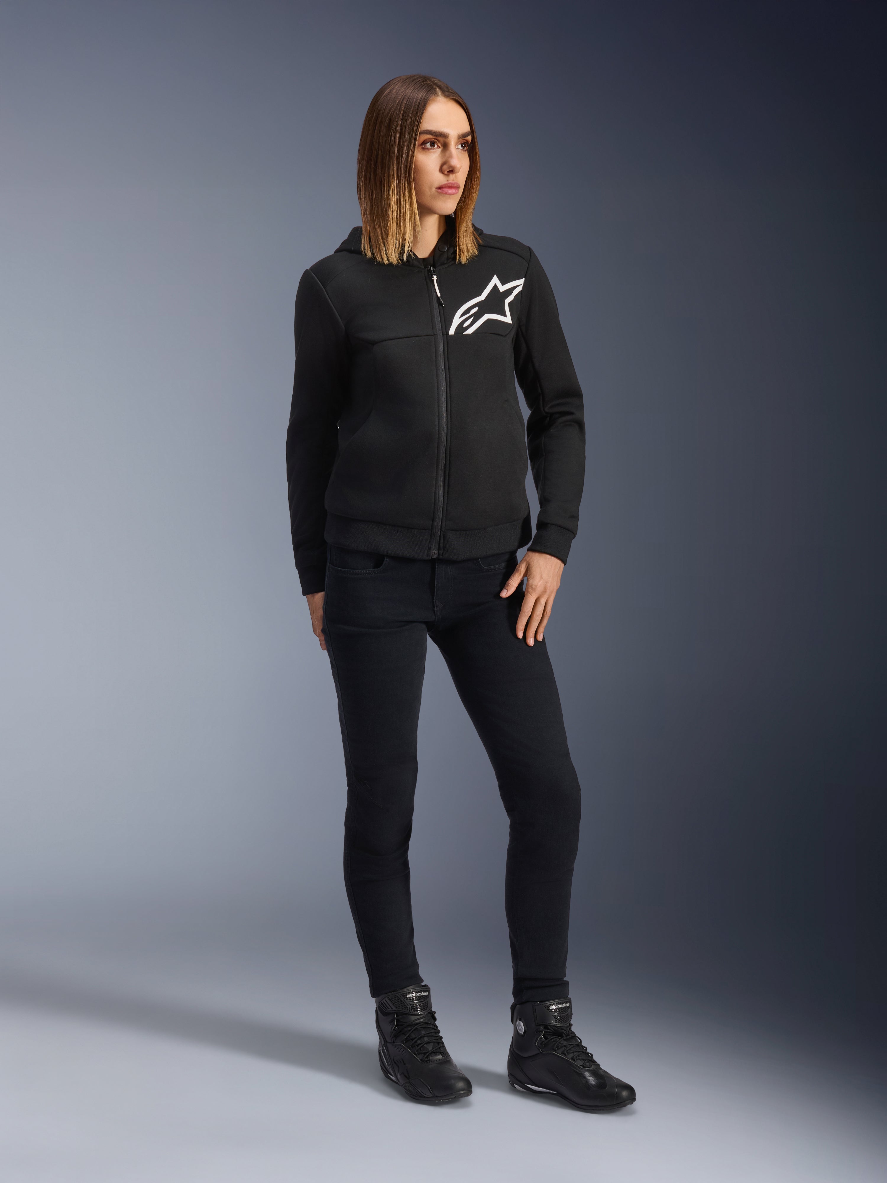 Mujer con Alpinestars Daisy V3 Women's Riding Denim, pantalones de moto para mujer en color Rinse Black, corte ajustado, combinados con sudadera negra con capucha y botas de moto, equipamiento de protección urbano casual